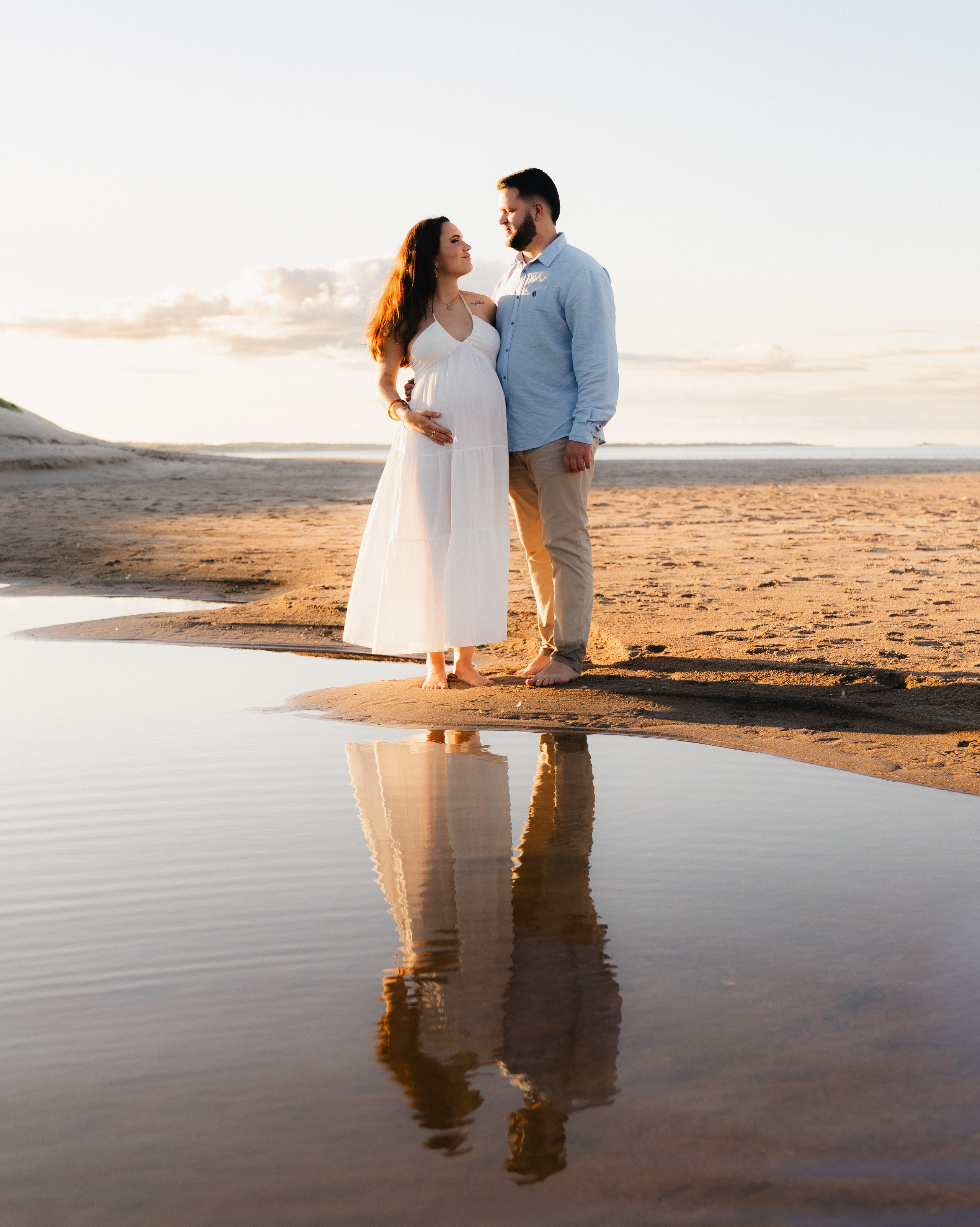 Couple during a golden hour maternity photoshoot at Druridge Bay in Northumberland, standing on the beach with reflections in the water.
