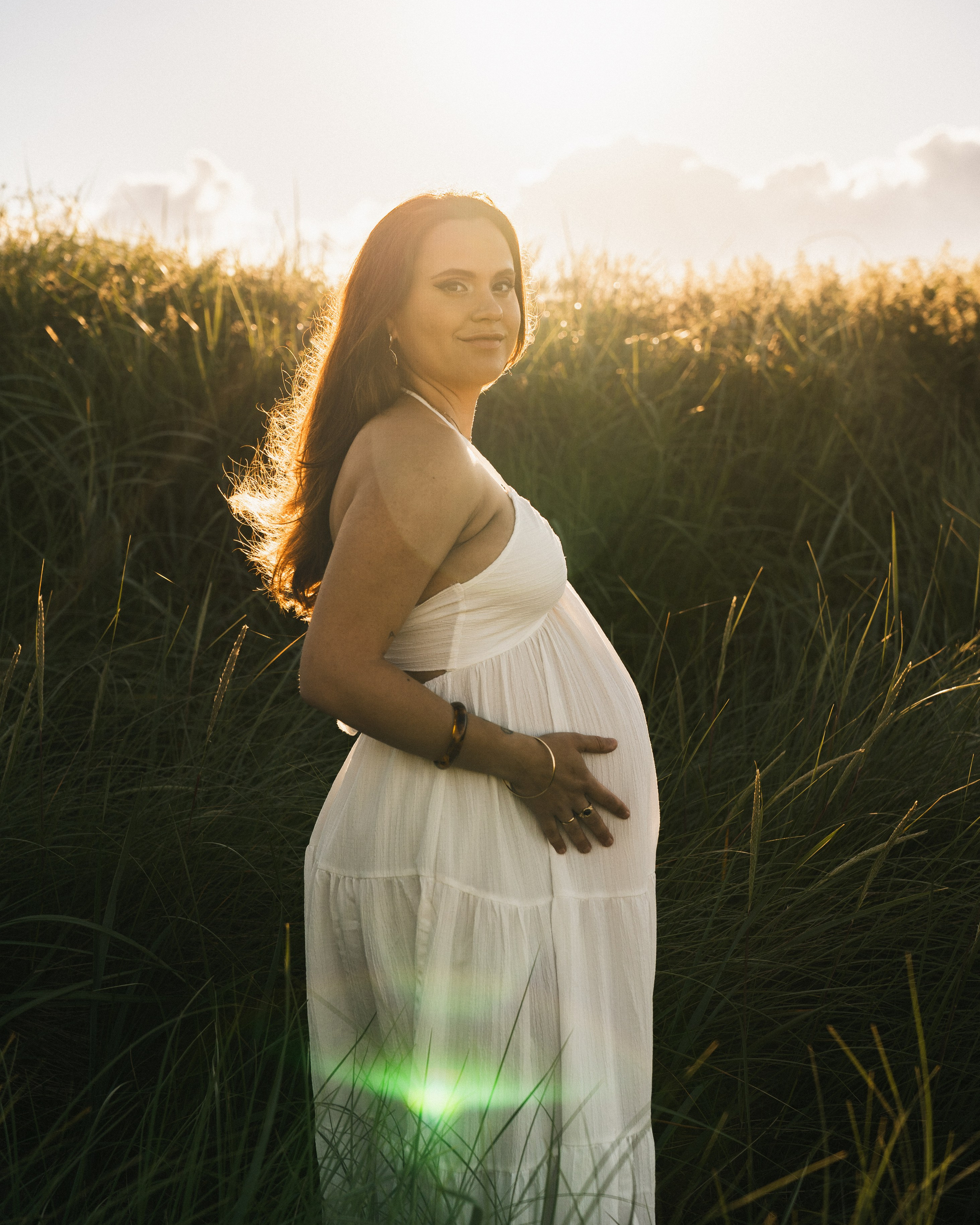 Pregnant woman in a white dress during a golden hour maternity photoshoot at Druridge Bay, in Northumberland, standing in coastal grass with sunset backlight. i