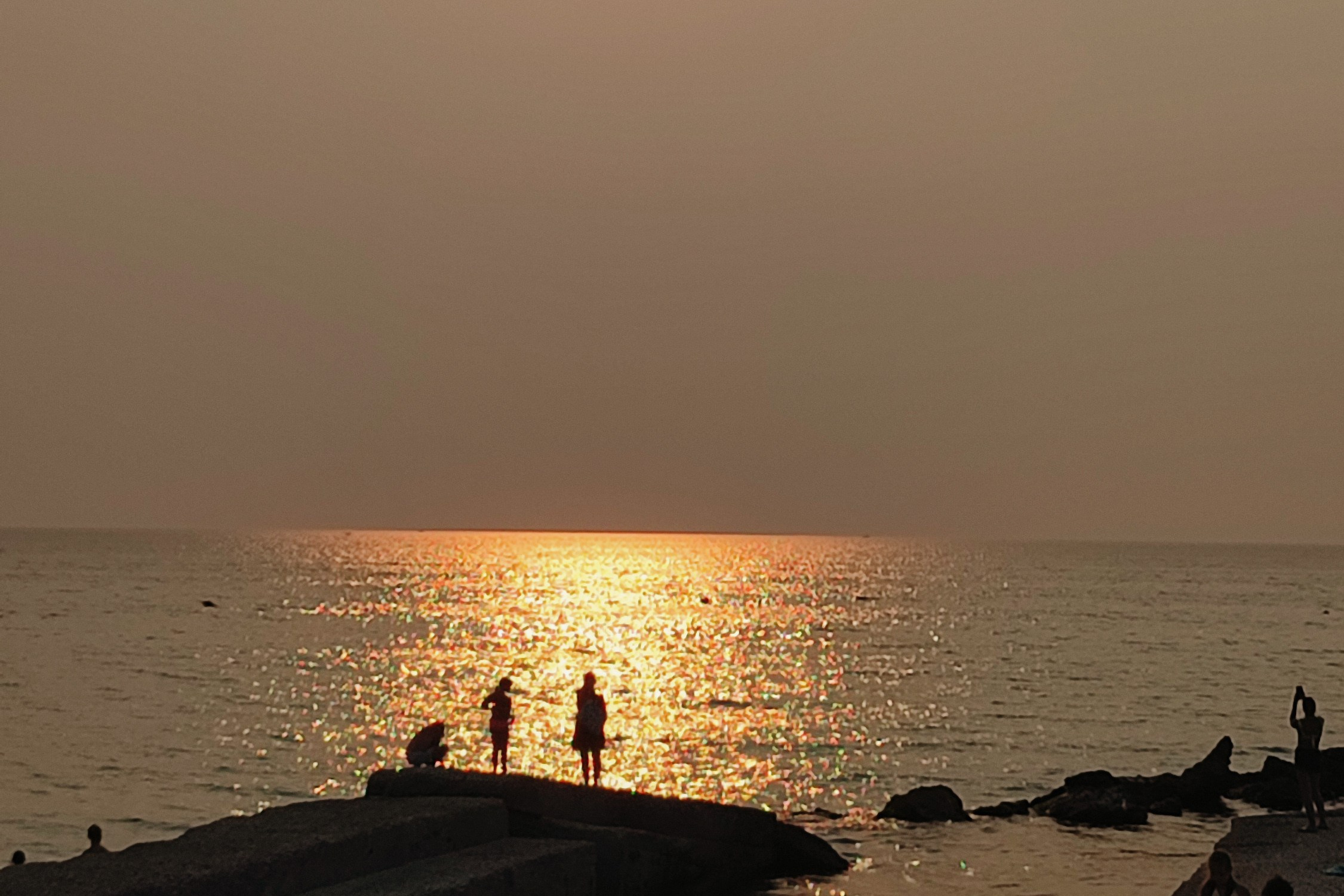 The sun sets over the sea, casting golden light across the water and the concrete piers where silhouettes of people are walking. The mood of a warm summer evening in Salento.