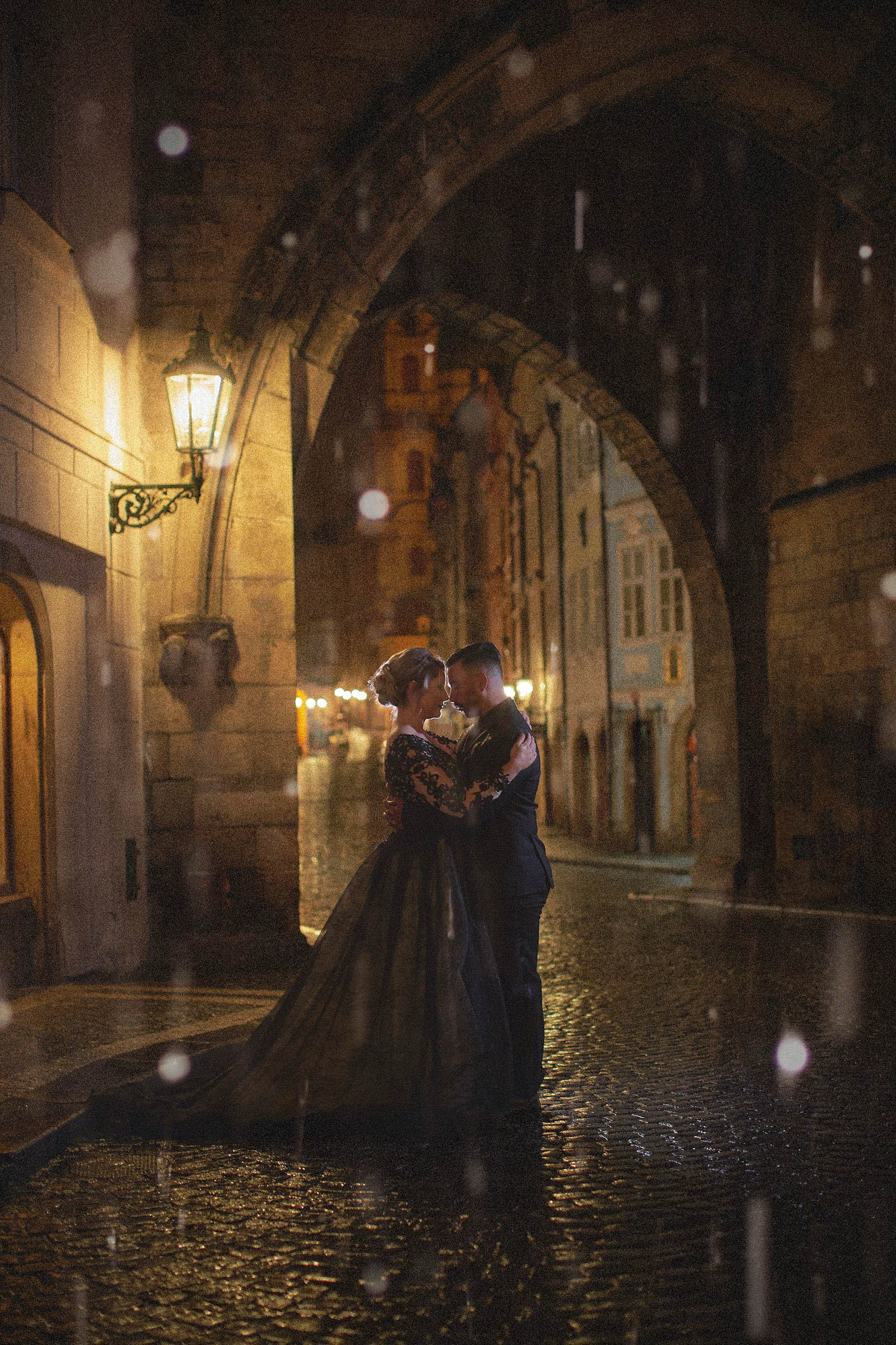Gothic couple in black embracing pouring rain nighttime Charles Bridge Prague anniversary.