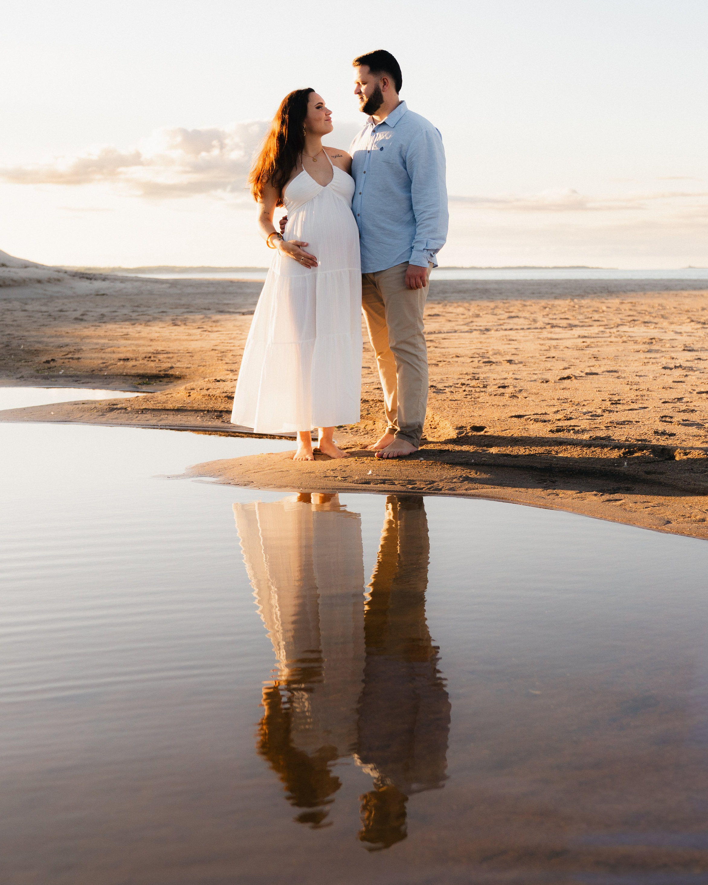 Pregnant couple standing together at sunset on Druridge Bay beach, Northumberland. Their reflection shows in shallow tidal water with golden light and dunes in the background. Maternity photography by Yana Balatskaya, Newcastle.