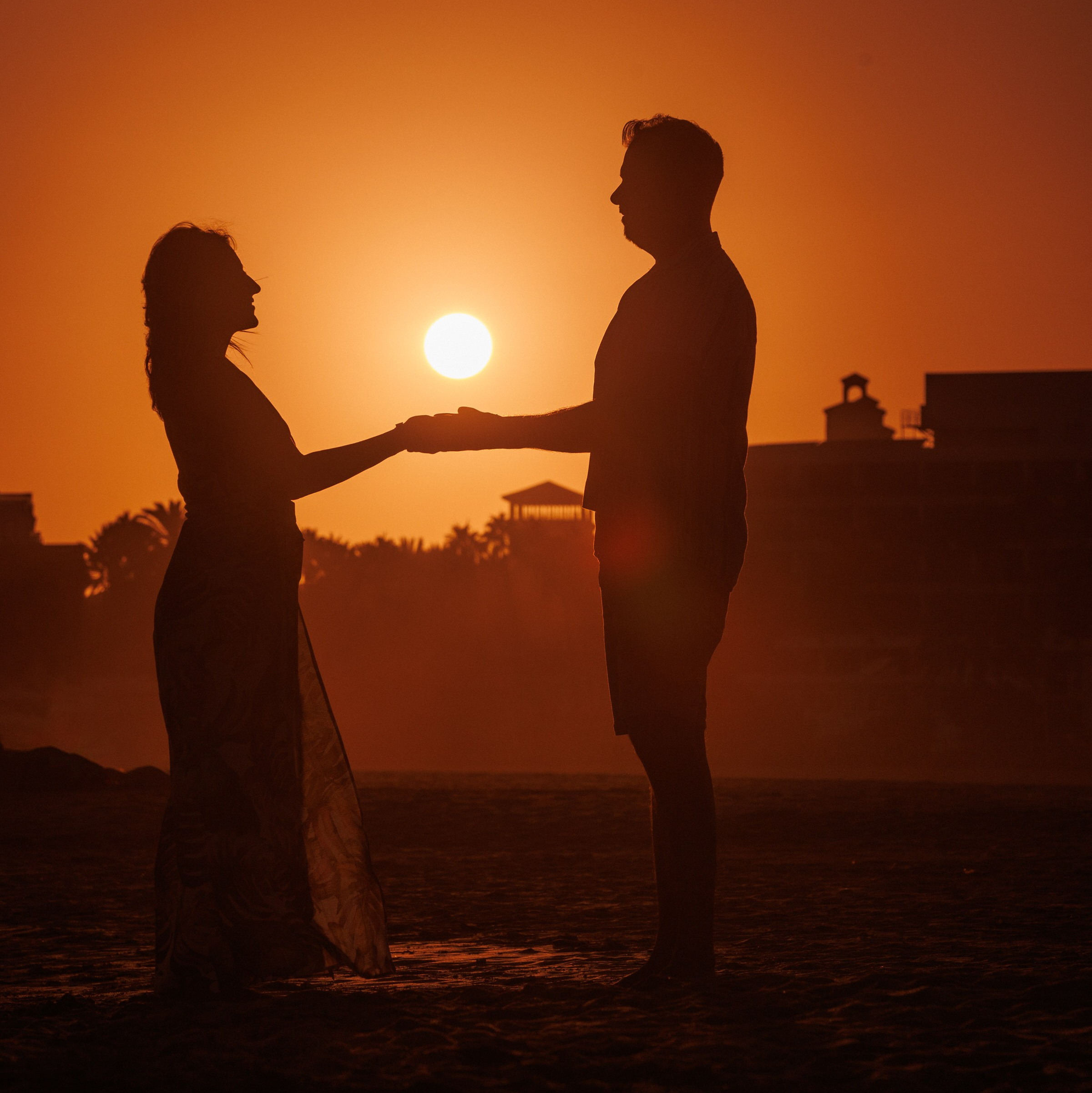 Silhouette of a couple holding hands on a beach at sunset, with the sun low in the orange sky and buildings in the background. Photographer in Maspalomas Beach