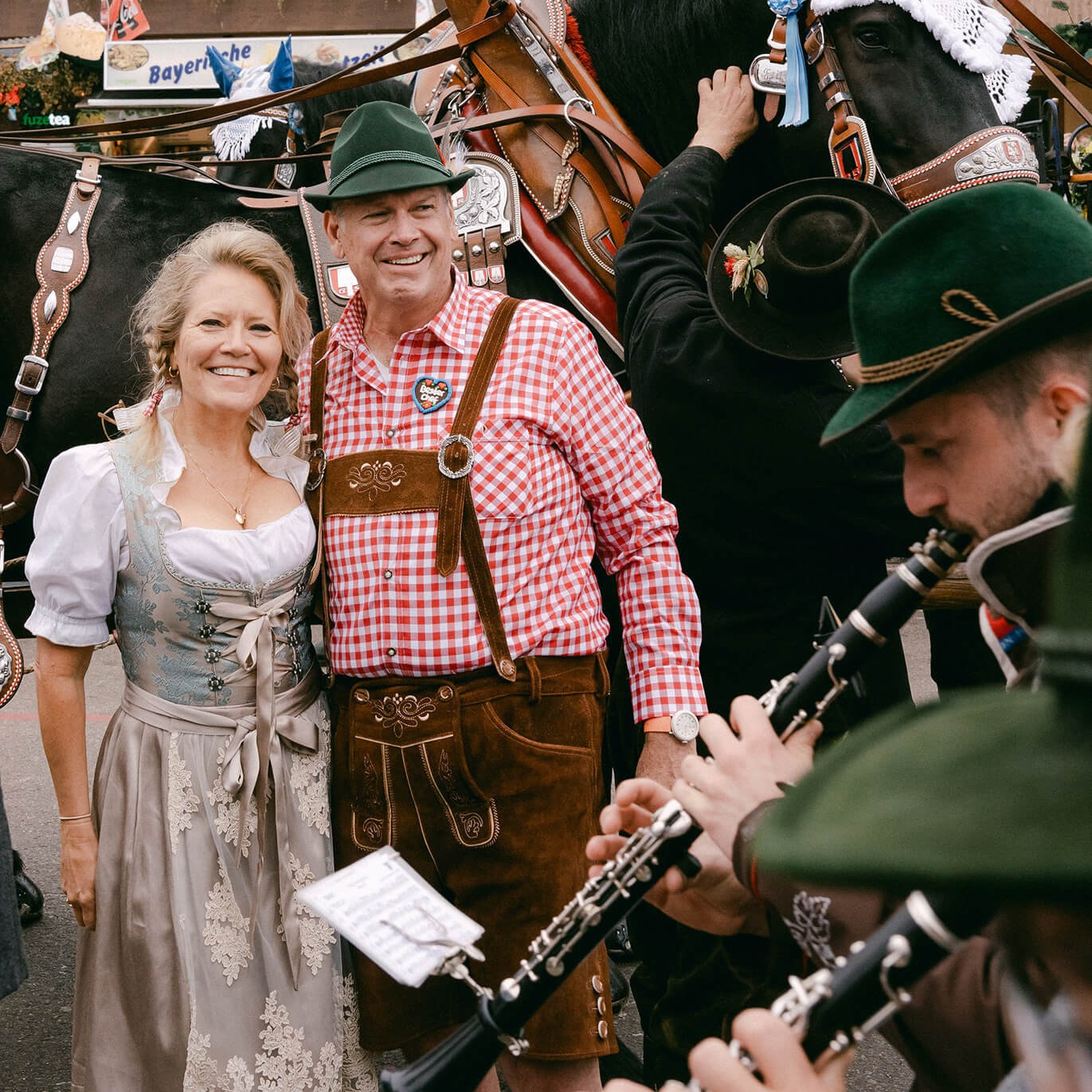 Paar im Dirndl und in Lederhose lächelt neben einer Pferdekutsche und traditionellen Musikern auf dem Oktoberfest in München