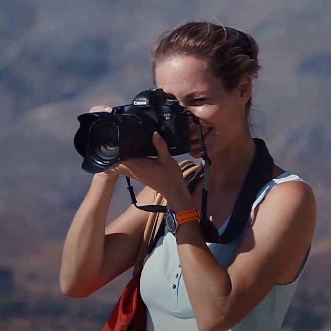 Фотограф Екатерина Романова, снимает с камерой в руке на фоне гор в Греции на Крите.