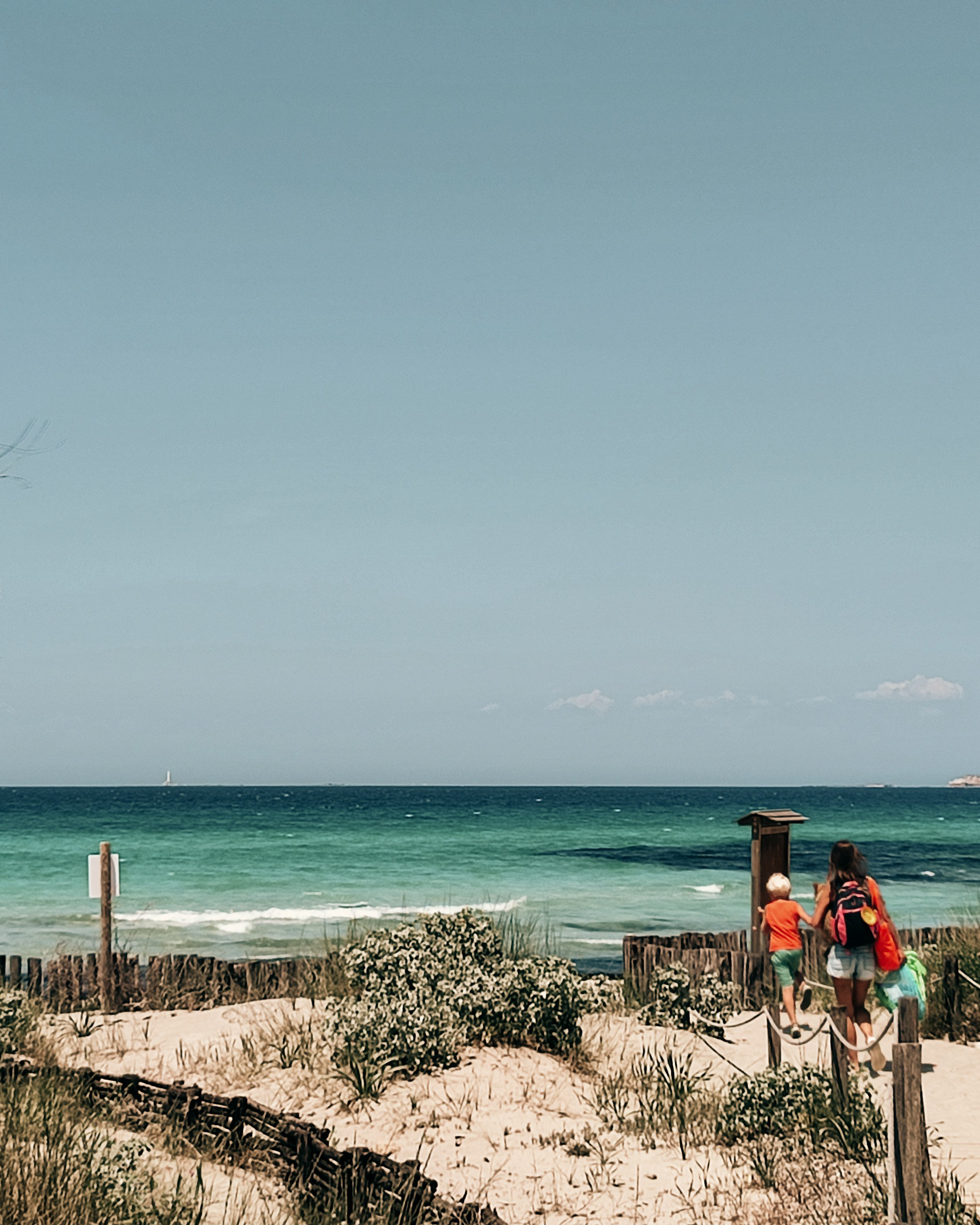A mother with children walks along a wooden path through sand dunes toward the turquoise sea. A summer day on one of Salento’s natural beaches.