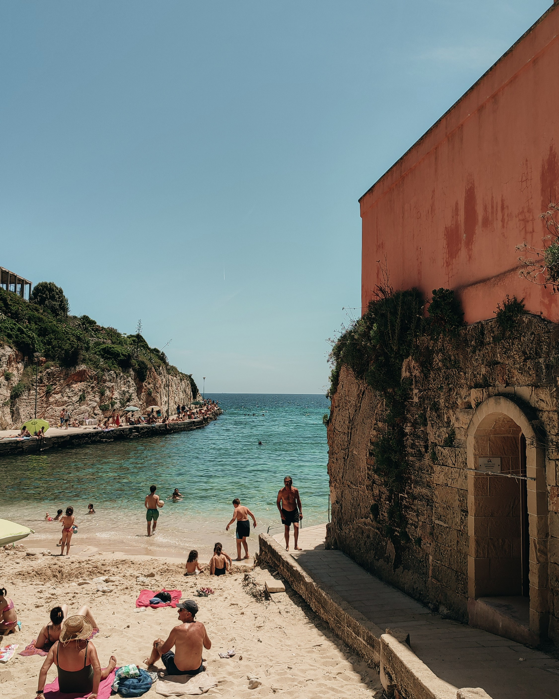 A sandy beach at the foot of an old wall in Tricase Porto. People relax in the shade of buildings, enjoying the view of the clear sea.