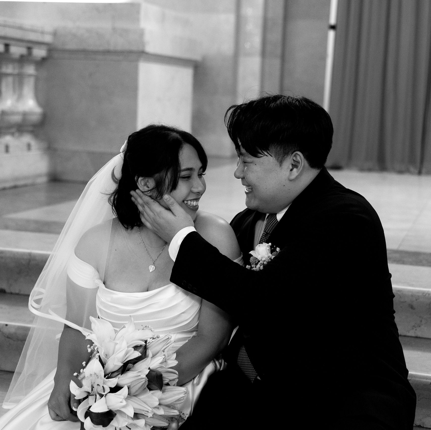 Black and white photo of a couple sitting on City Hall steps, groom touching bride’s face