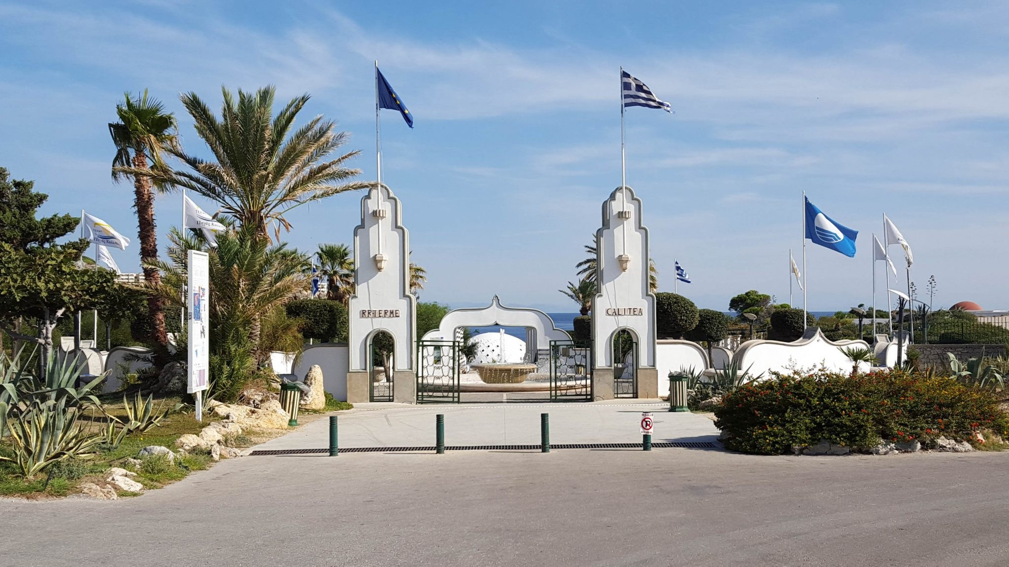 Sehenswürdigkeit auf Rhodos: Eingang zu den Kallithea Quellen (Kallithea Springs) mit historischer Architektur, Palmen und Flaggen. Beliebte Hochzeitslocation und Ausflugsziel in Griechenland am Mittelmeer.
