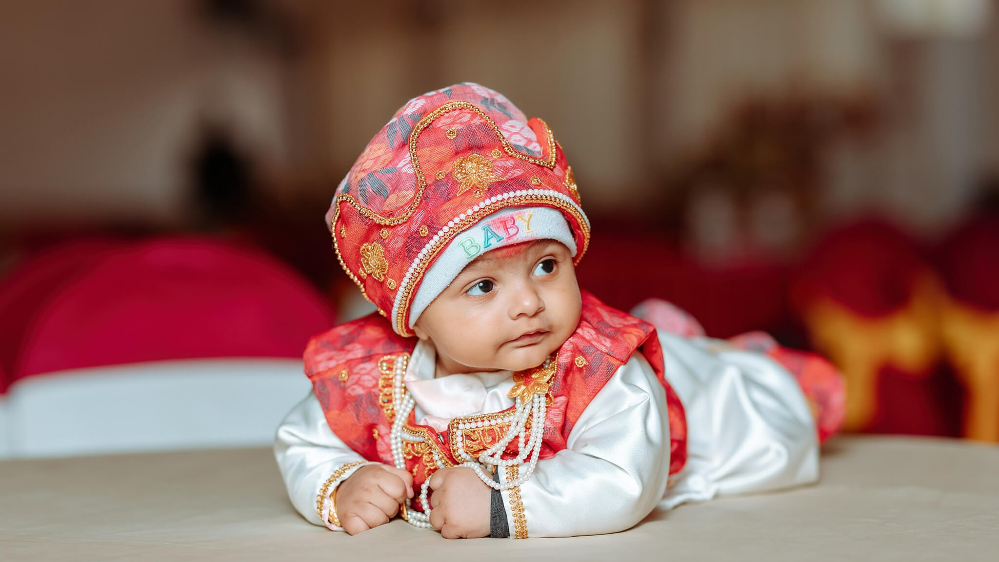 Baby lying on its tummy wearing a red embroidered cap and outfit during a Pasni baby photoshoot