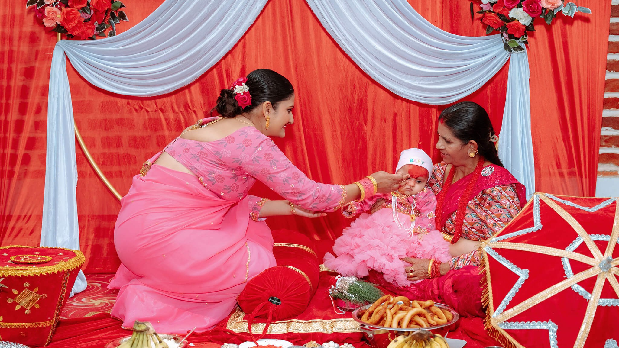 Family performing the Pasni rice feeding ritual in front of a red and white draped backdrop in Butwal
