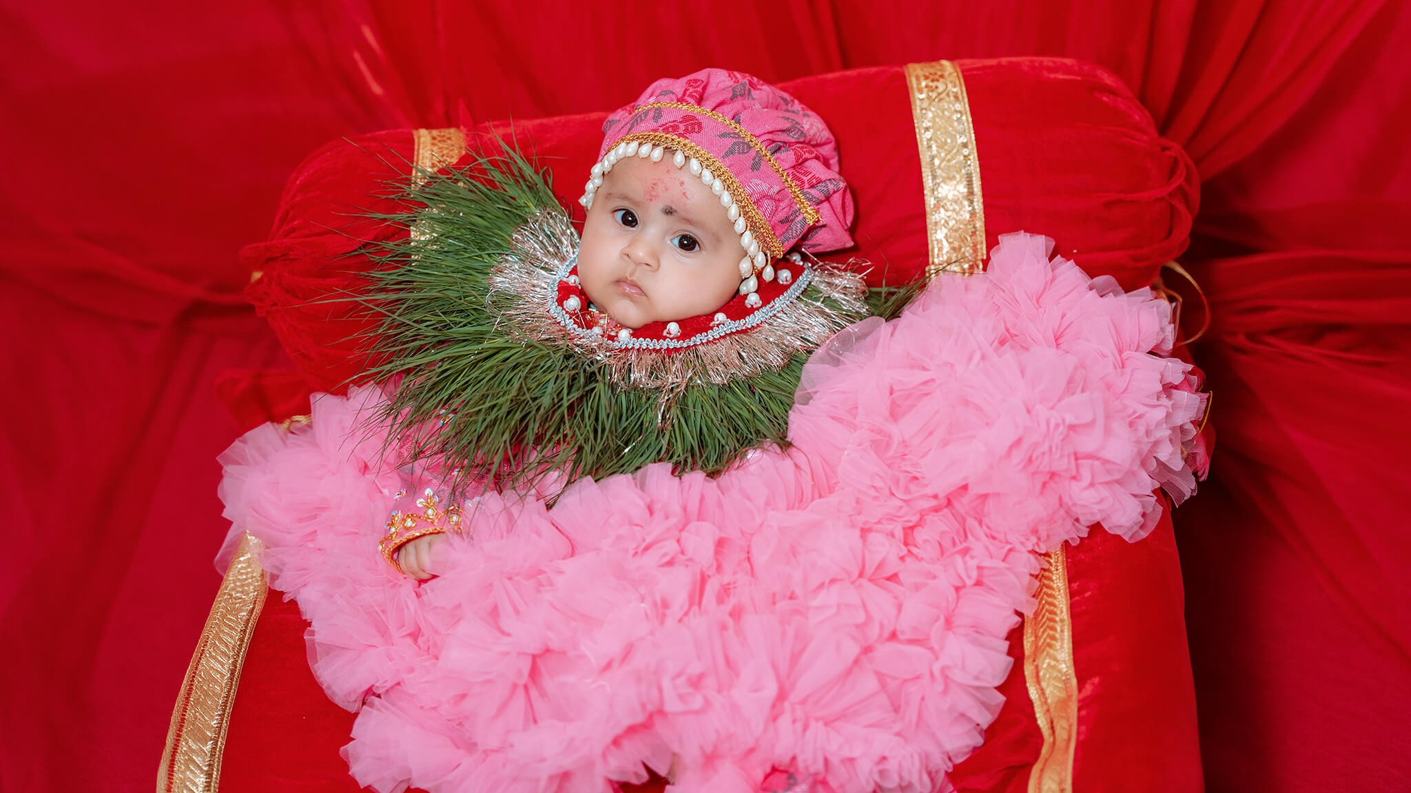 Baby in a red outfit with a pink feather trim and floral garland held gently by parents