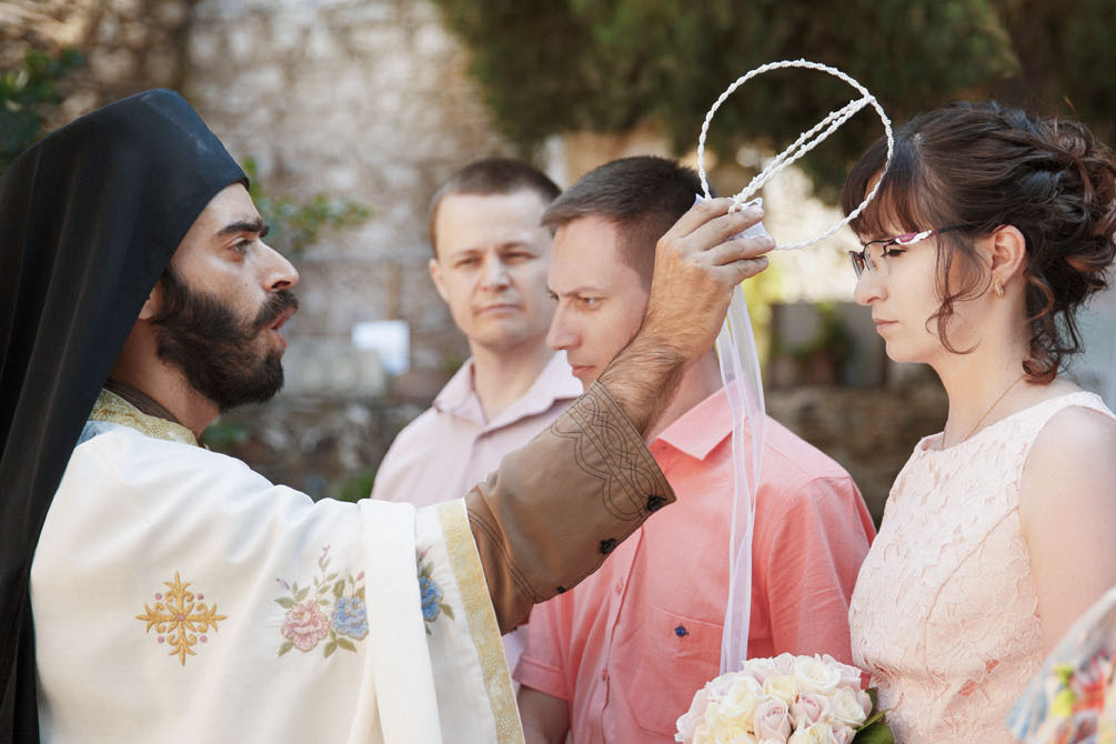 Святой отец на венчании держит венки над головой невесты. Фотограф на Крите, свадьба в Греции.