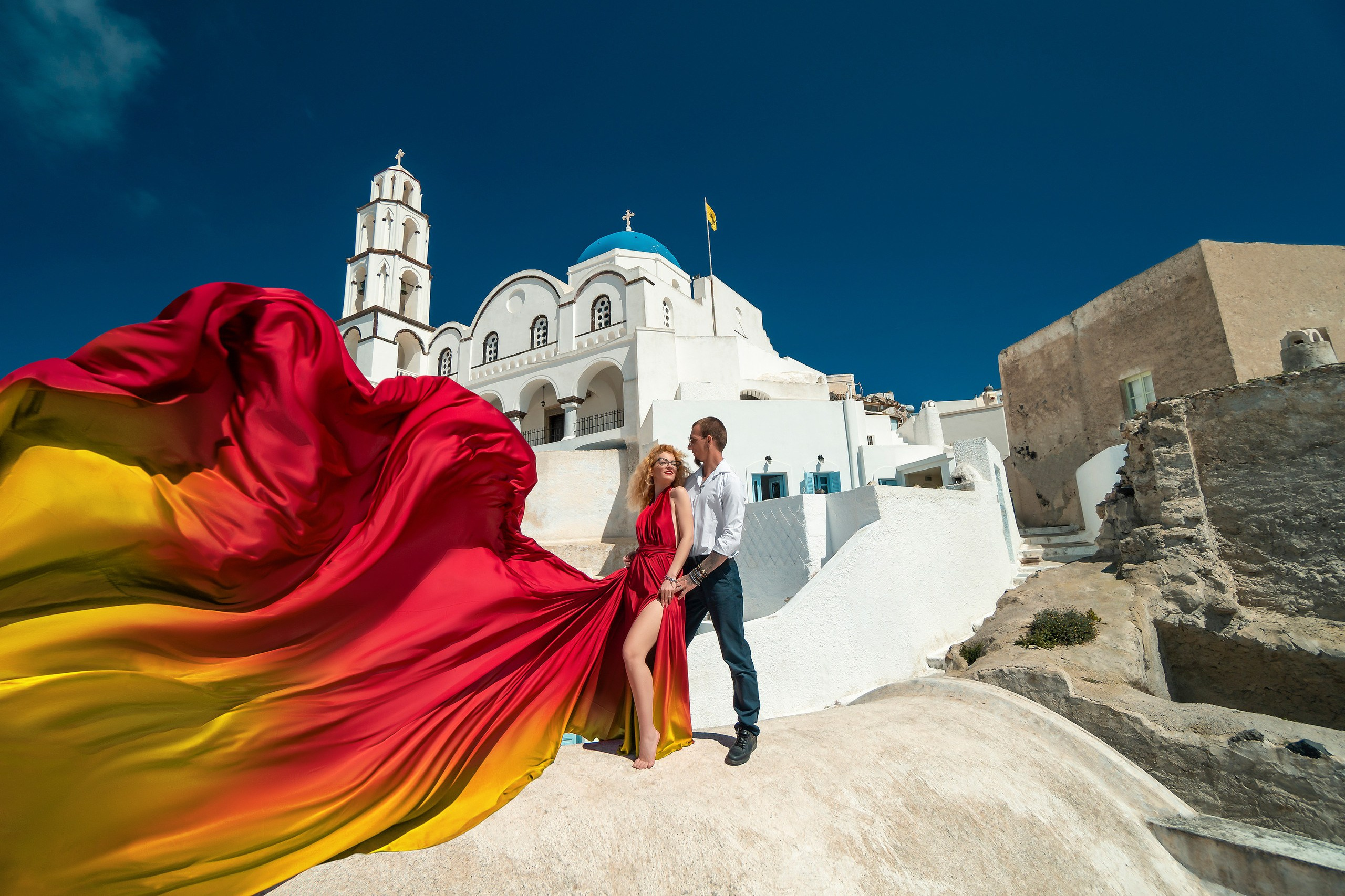 A captivating photoshoot featuring a woman posing in a flowing flying dress, captured against the stunning whitewashed buildings and blue-domed churches of Santorini, Greece. The image highlights dynamic poses that showcase the dress's movement, set against the picturesque Aegean Sea backdrop, ideal for fashion and travel photography enthusiasts.
