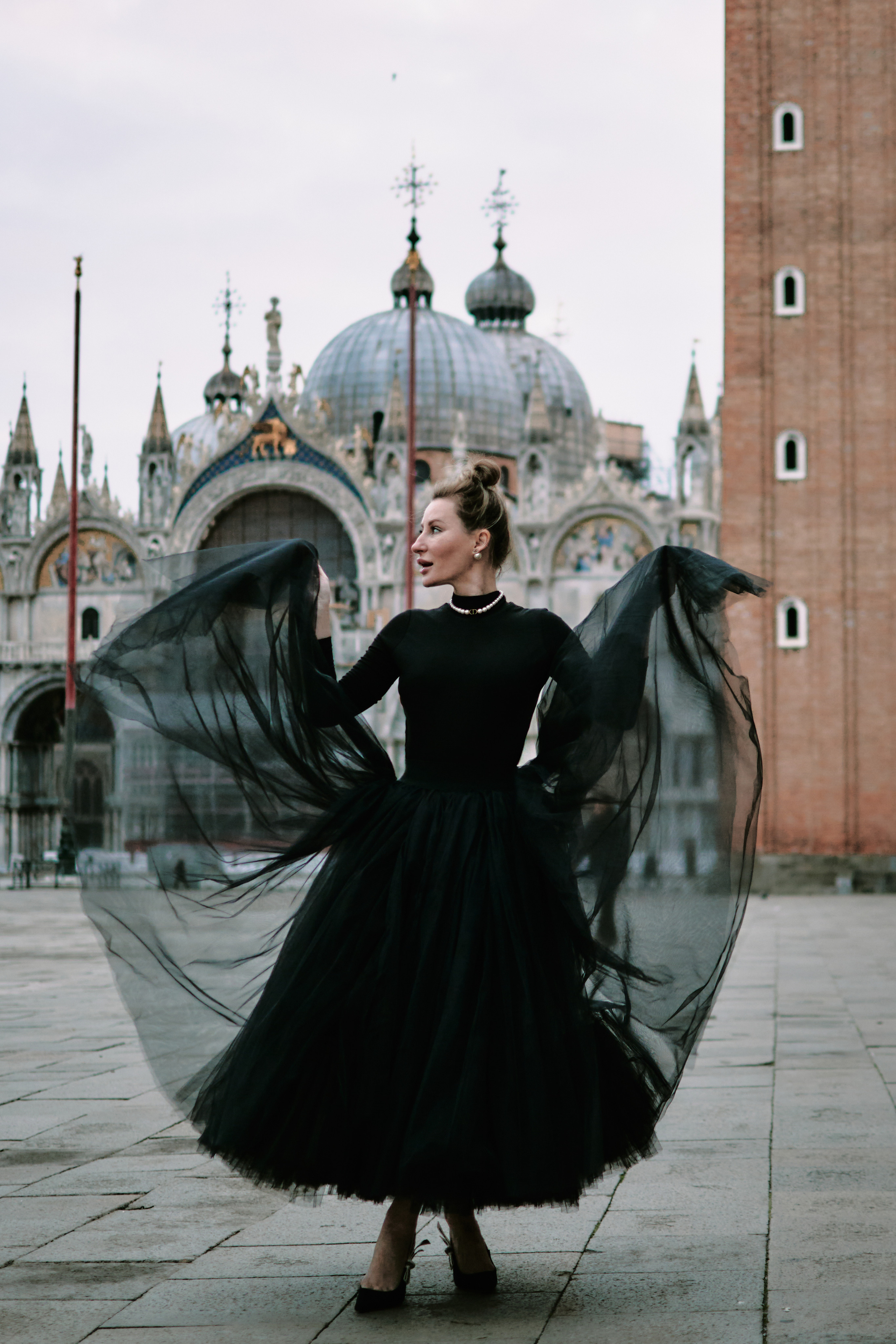 A young woman stands on the crowded San Marco Square in Venice. She wears a black flowing dress and looks away from the camera, her hair blowing in the wind. The historic buildings and the iconic Campanile tower rise behind her, creating a stunning backdrop. Tourists and pigeons are visible in the background, adding to the lively atmosphere of the square. The photo captures the beauty and charm of Venice, making it a perfect representation of this romantic city. In the background, the iconic St. Mark's Basilica stands tall, its intricate architecture and golden mosaics glistening in the sunlight. The bustling square is filled with people and the sounds of music and laughter, creating a lively and romantic atmosphere. Fashion blogger shoot Venice. Portrait and Fashion Photographer in Venice, Italy. Instagram Photos with lifestyle photographer