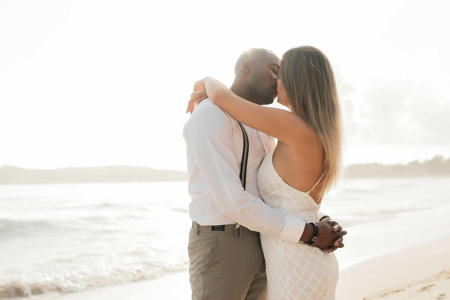 The newlyweds are kissing on the beach at dawn, the groom has a white shirt and the bride is wearing a classic wedding dress. 