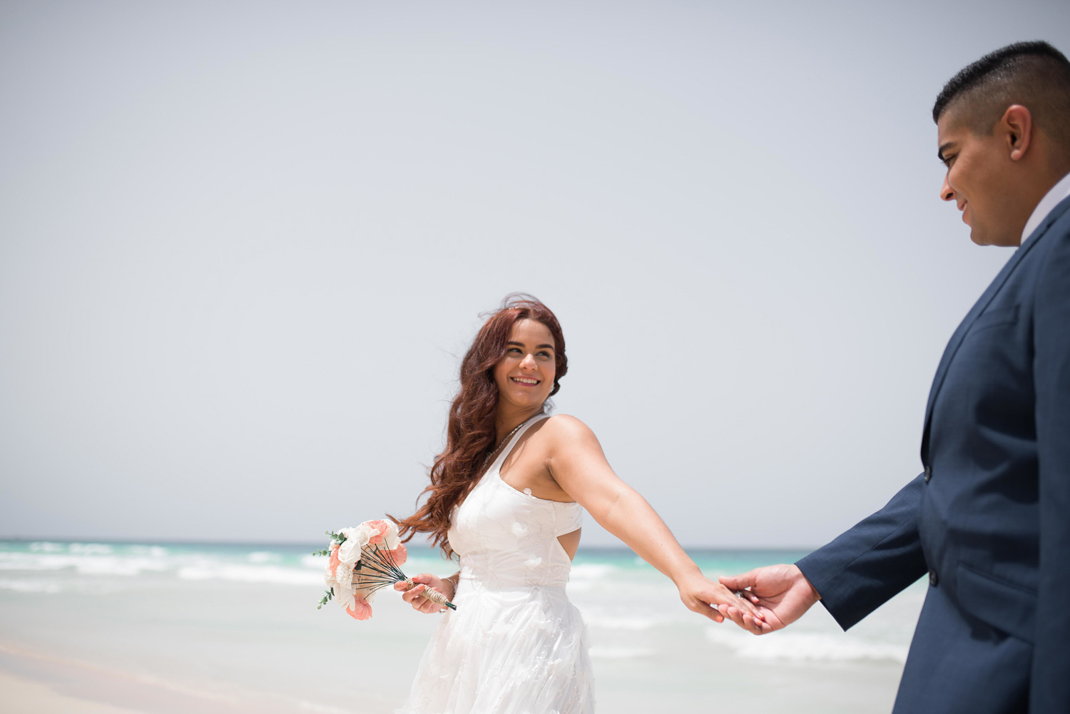 Жених держит за руку невесту, а невеста смотрит не жениха. Невеста держит букет в руке а жених одет в синий пиджак.