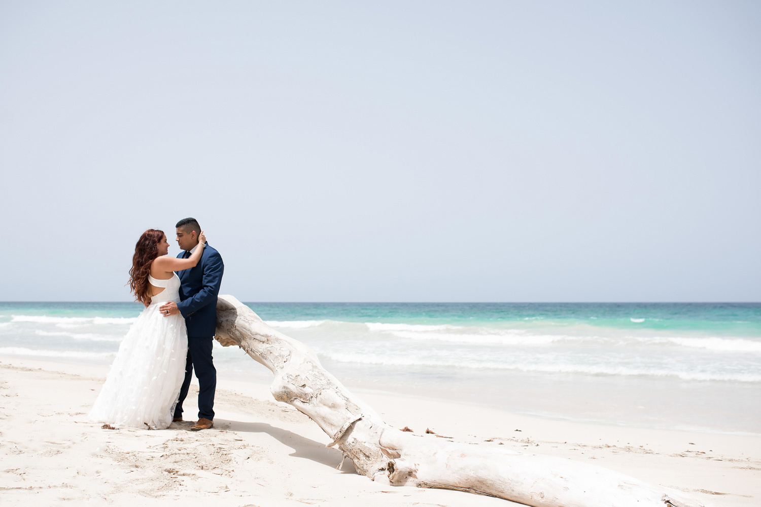 Невеста обнимает жениха на фоне океана, упершись в дерево которое вынесло на пляж вовремя шторма.