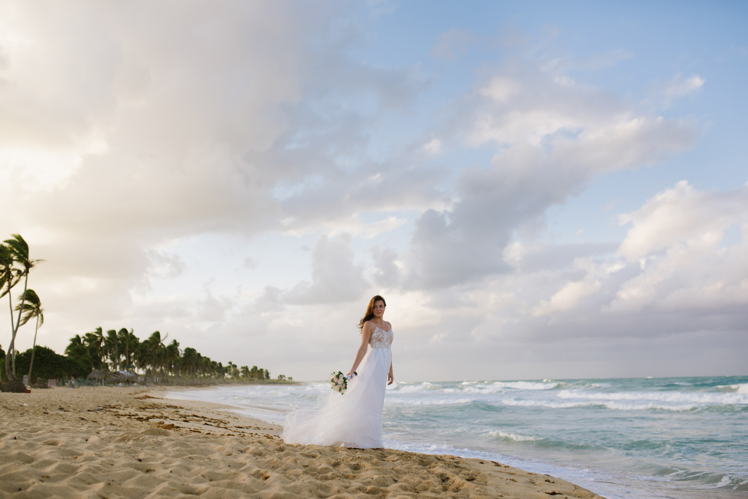 A bride poses for a photographer at sunset, she stands in a white dress against a backdrop of a sunset sky and ocean.