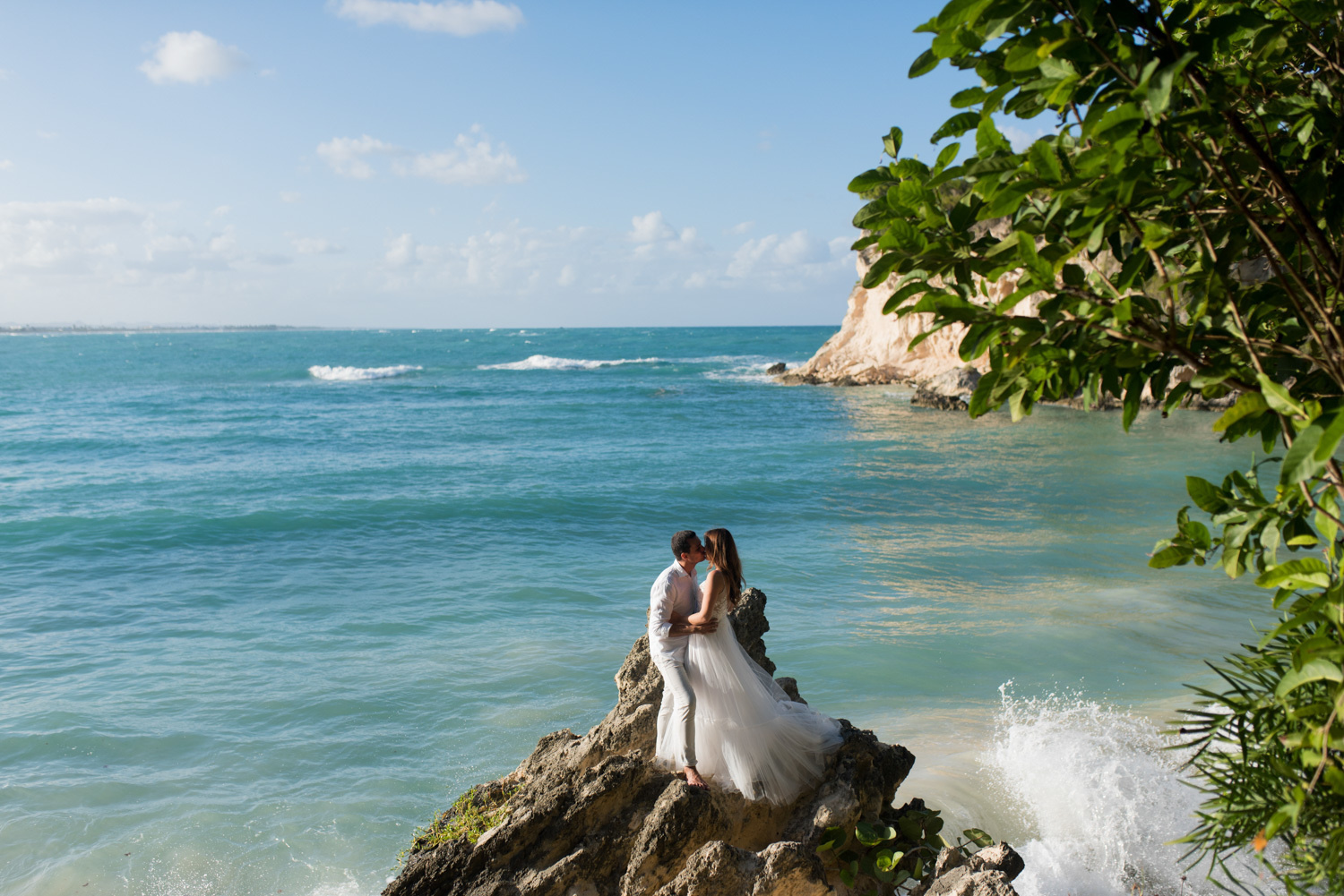 The bride and groom kiss on a rock, and the waves of the ocean cover the rock.