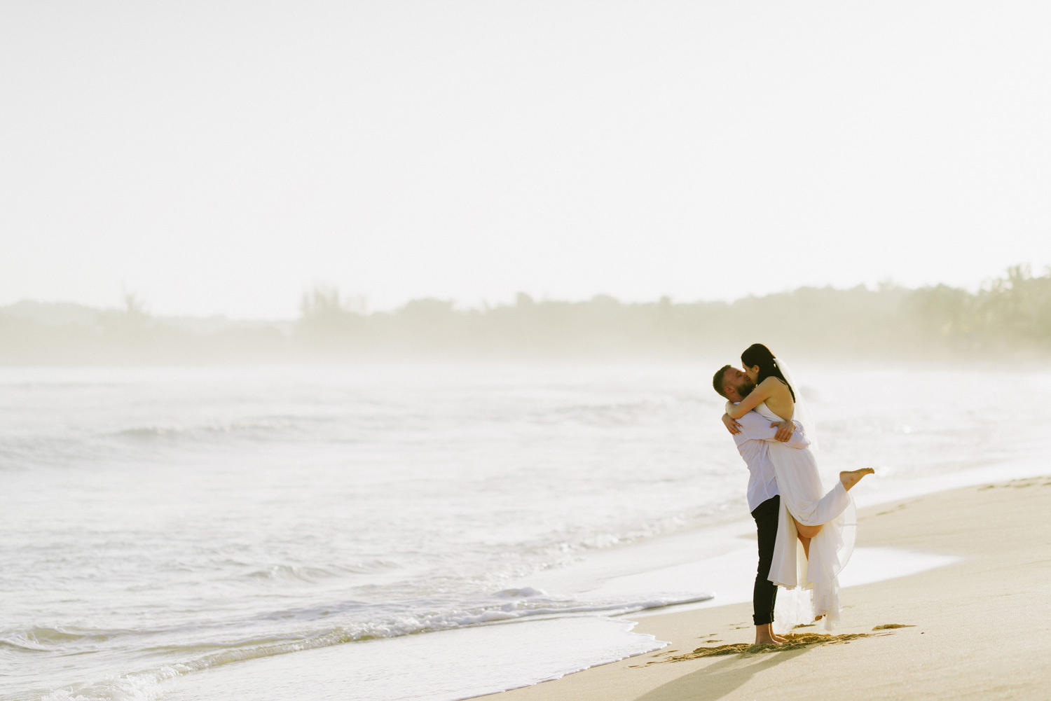 Жених поднял невесту на руки и целует ее на пляже Макао. 