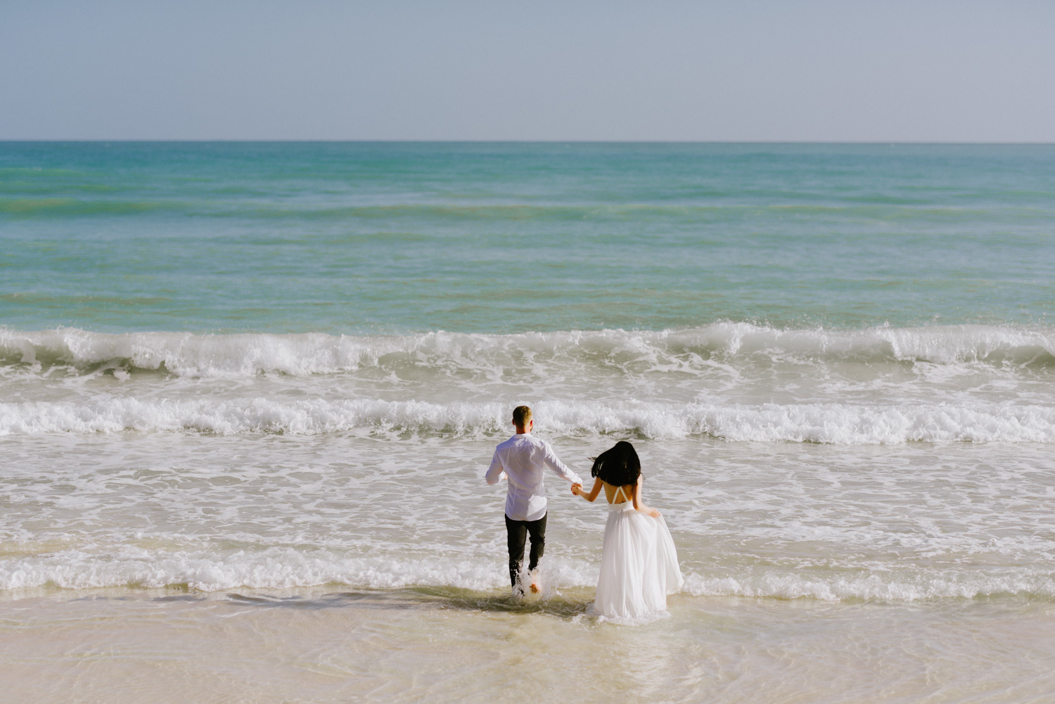 Невеста и жених бегут в океан держа друг друга за руки. 