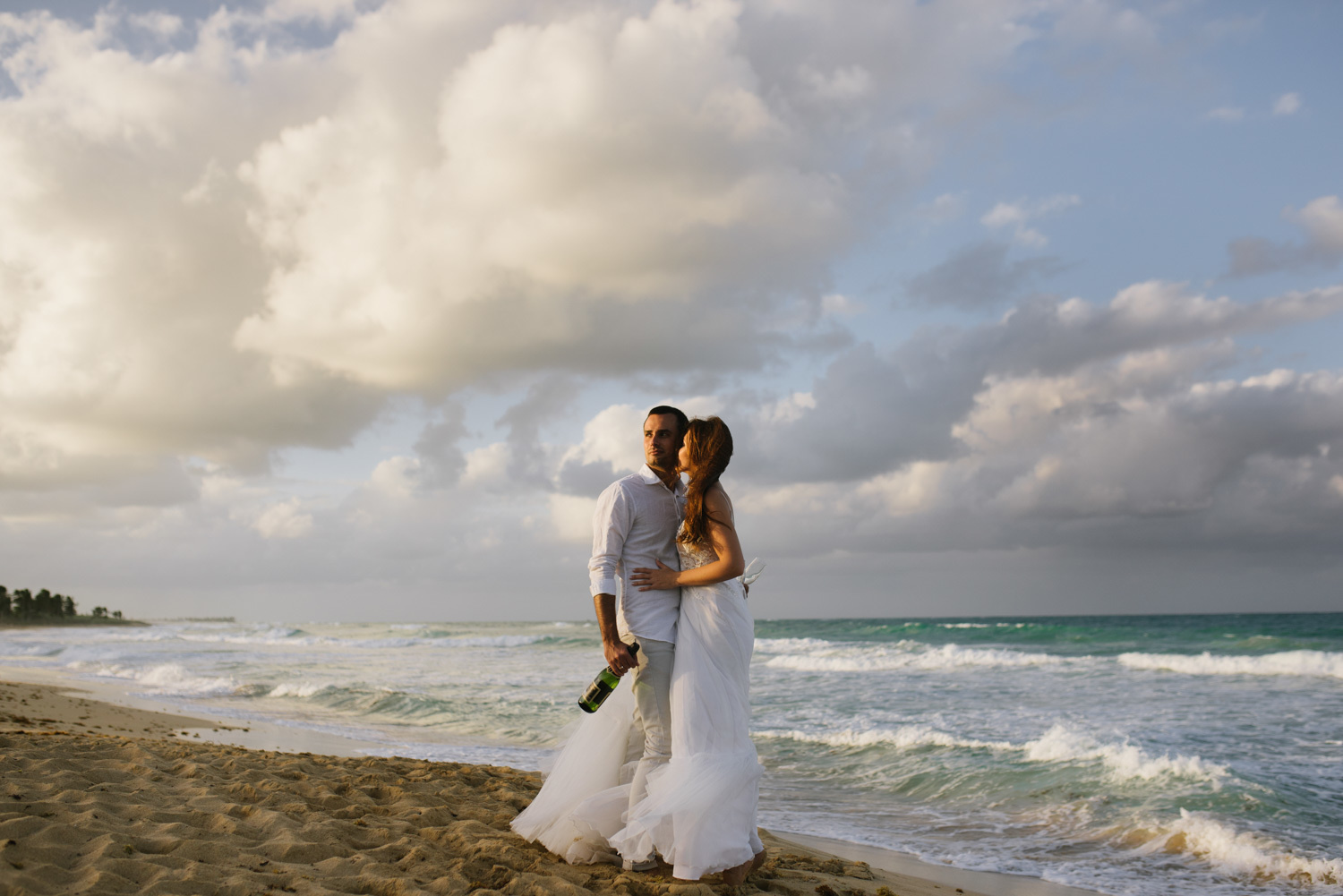 The newlyweds embrace each other against the ocean and the sunset sky, with the groom holding a bottle of champagne and the bride hugging him by the waist.