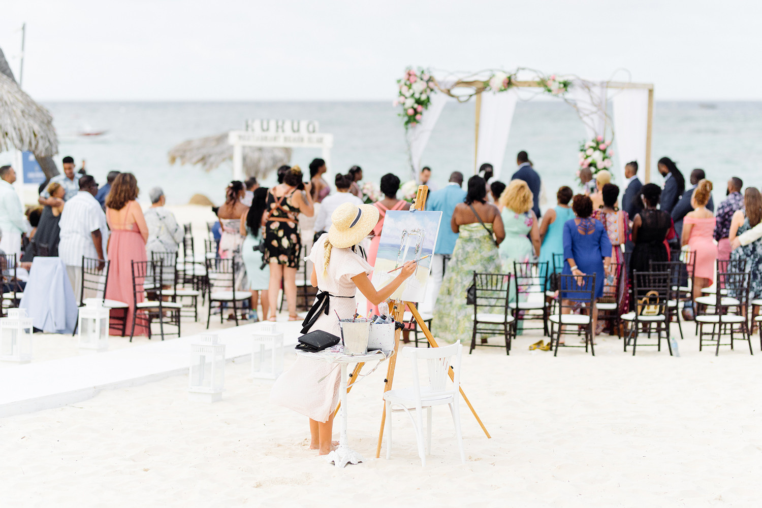 Художник рисует свадебную церемонию на пляже, она стоит спиной возле мольберта.