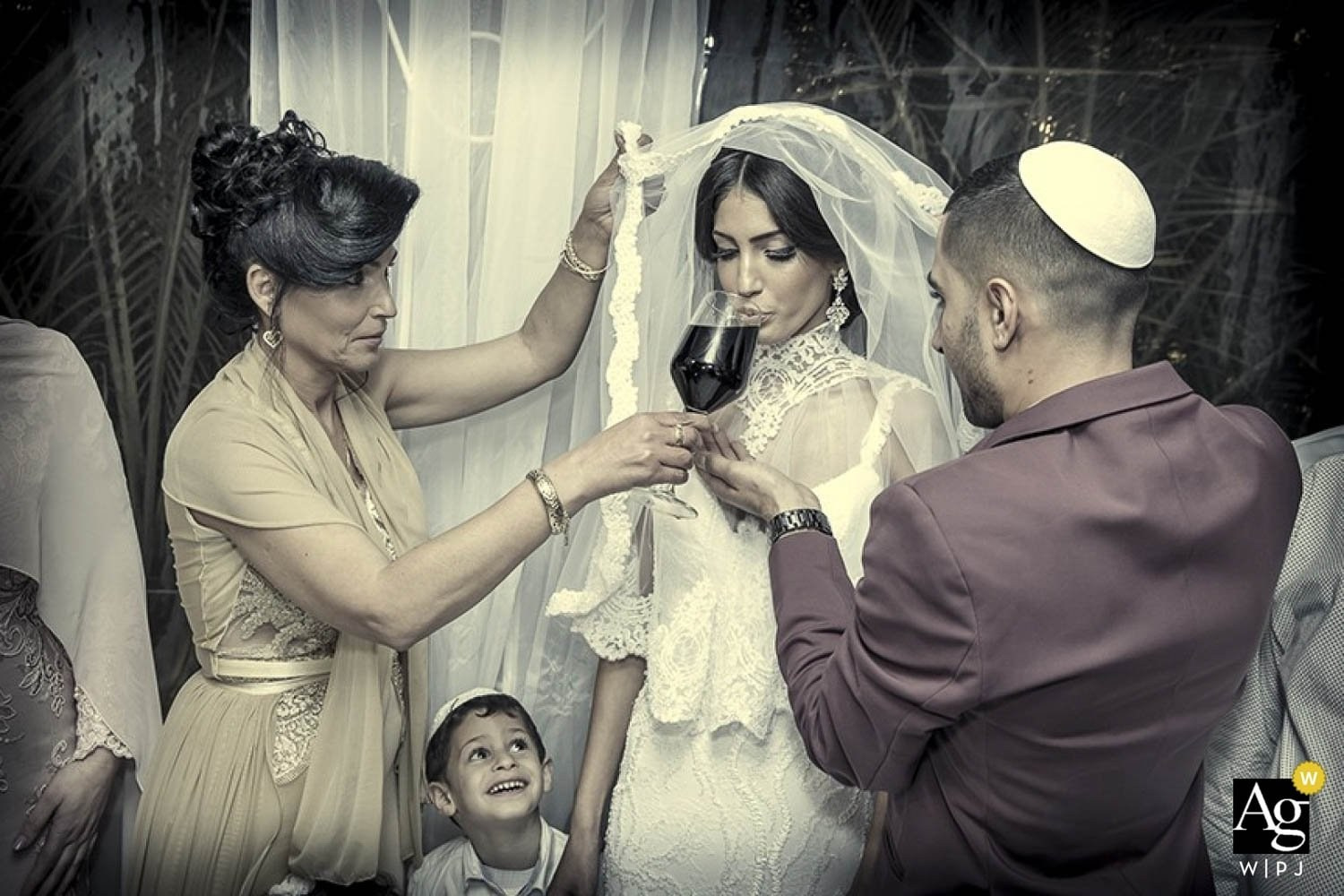 Jewish wedding ceremony: the bride drinks wine as the groom and a woman assist, with a smiling boy in the foreground
