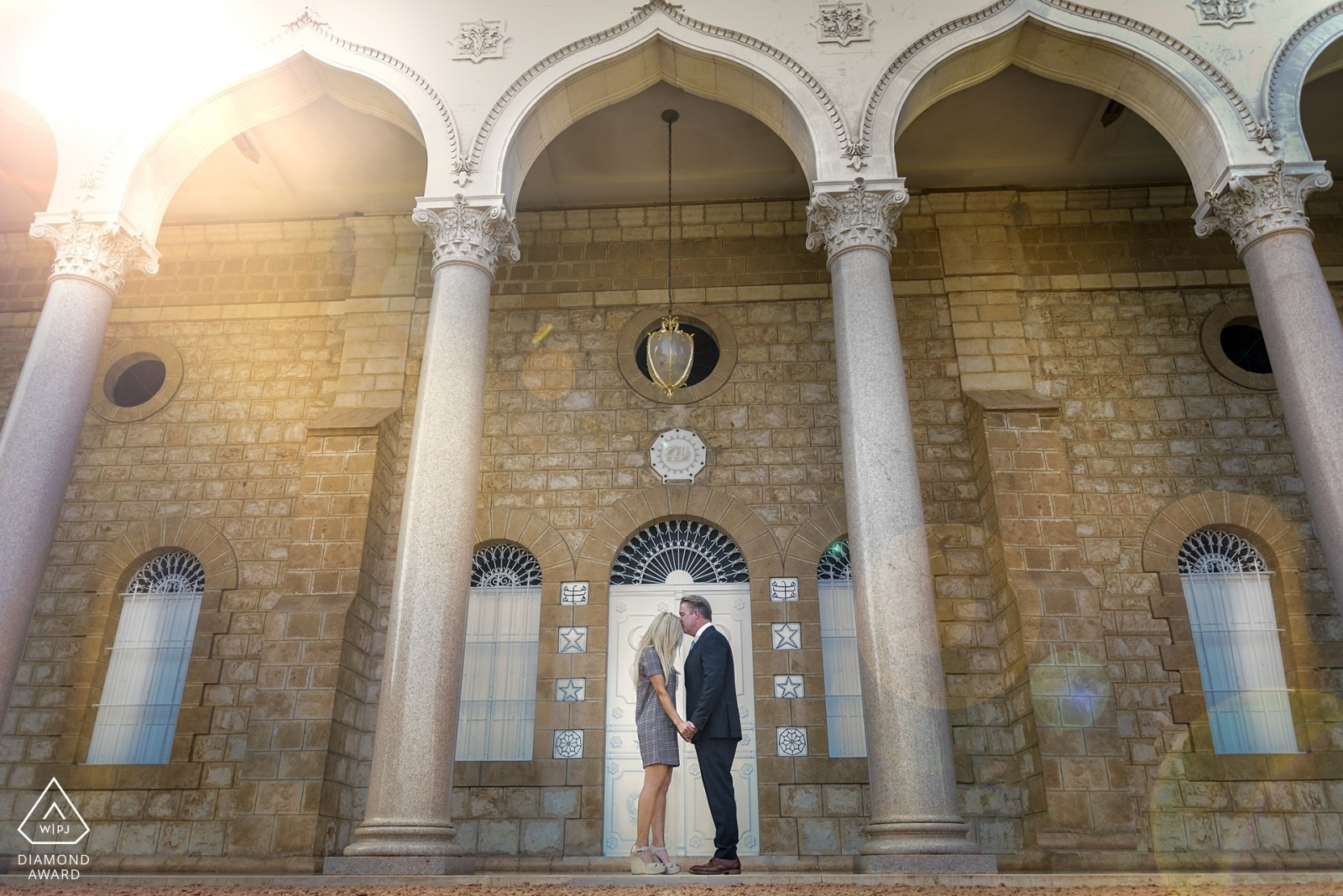 A couple stands in front of a majestic building with arches and columns in the Bahai Gardens in Haifa, holding hands, enjoying a romantic photo shoot against the backdrop of historical architecture on a sunny day