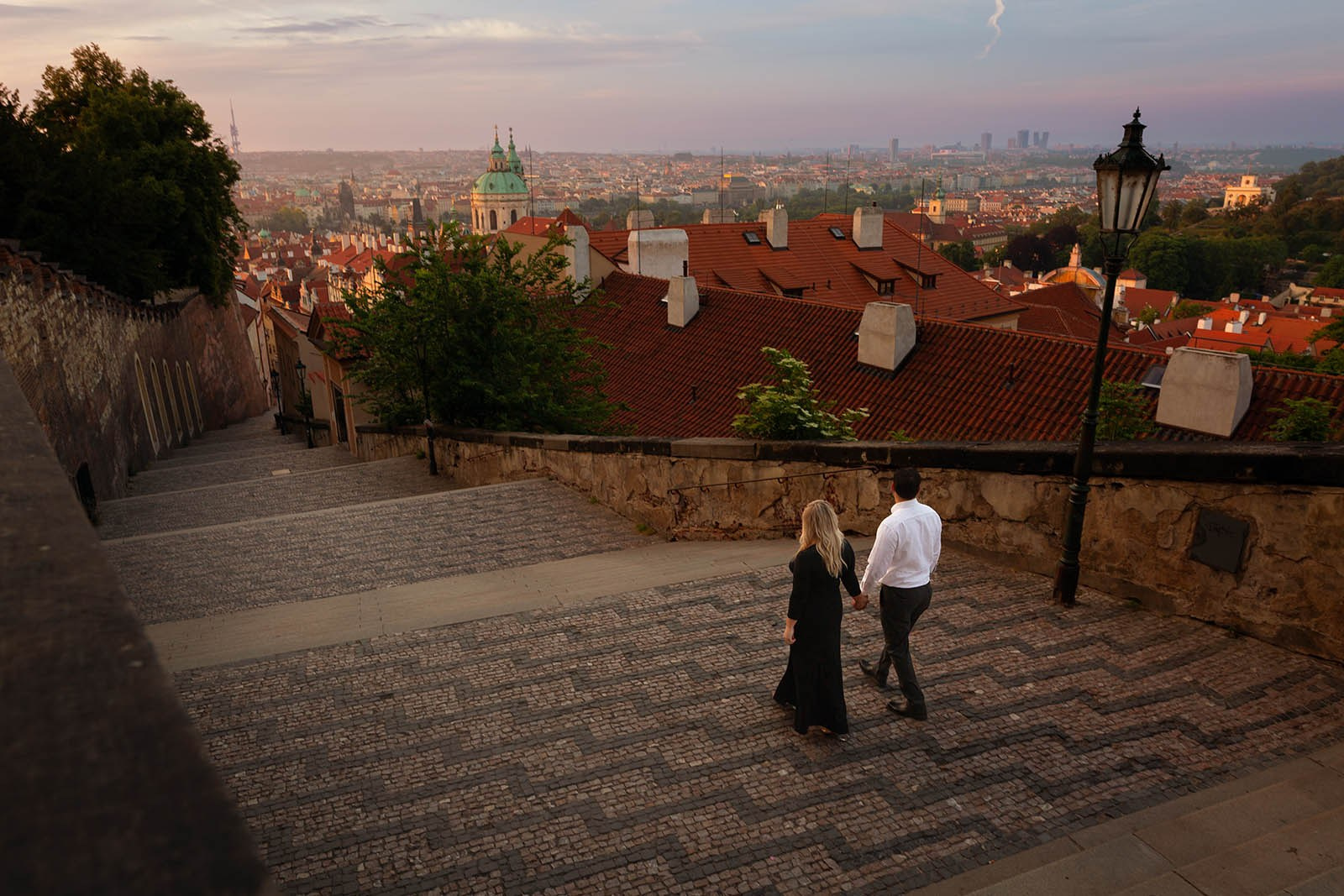 An American couple descending the castle steps toward Charles Bridge as sunrise light spreads across Prague