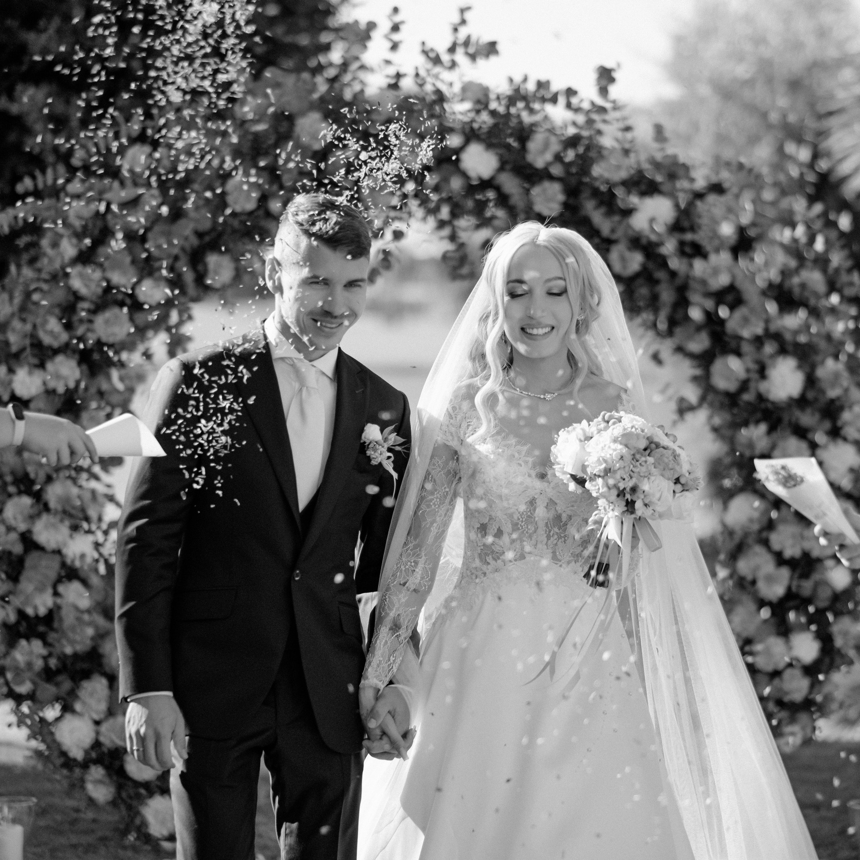 A newlywed couple walking down the aisle hand-in-hand, smiling as flower petals are thrown around them, with a floral arch and greenery in the background.