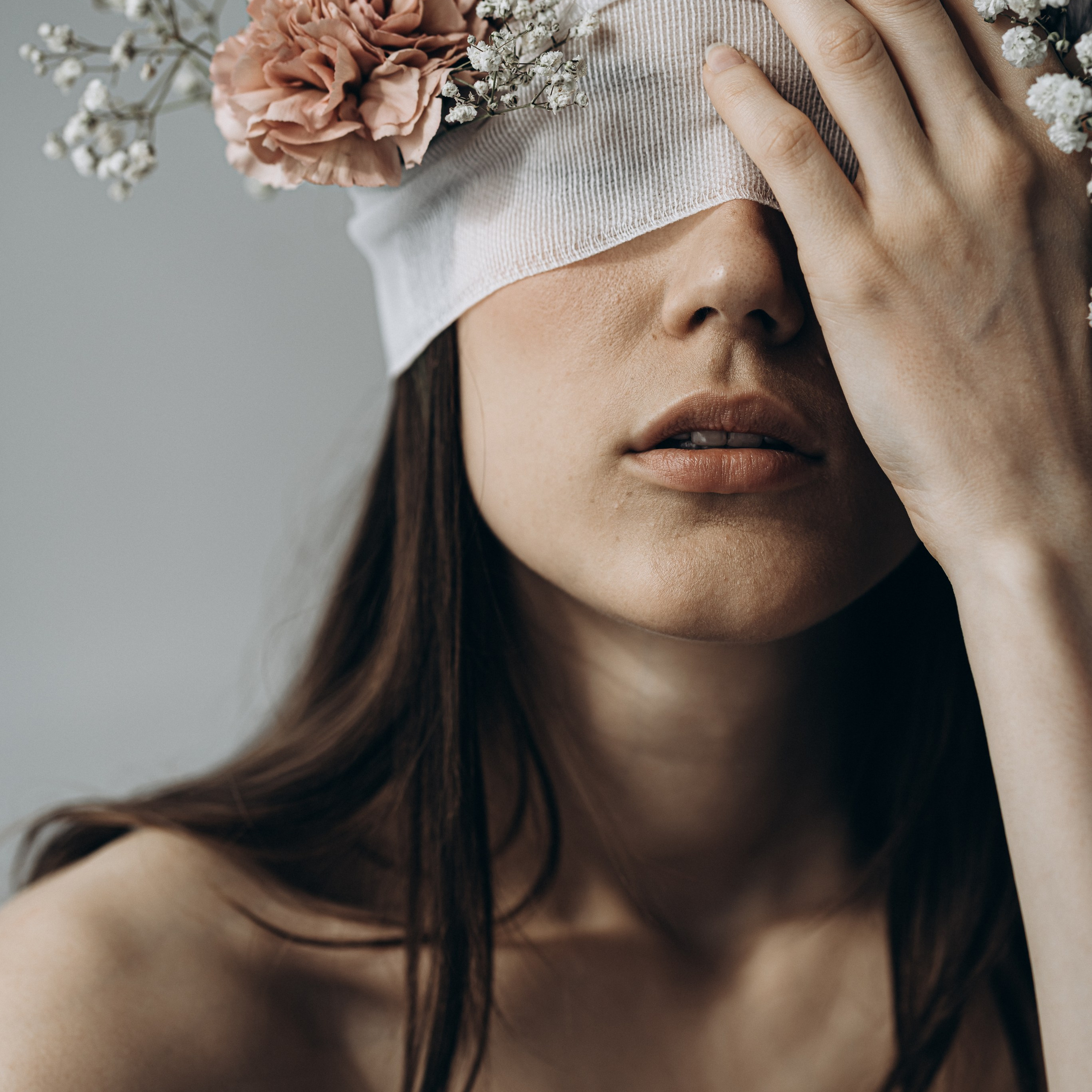 Colour portrait of a woman wearing a flower-covered blindfold, with one hand raised to her face.