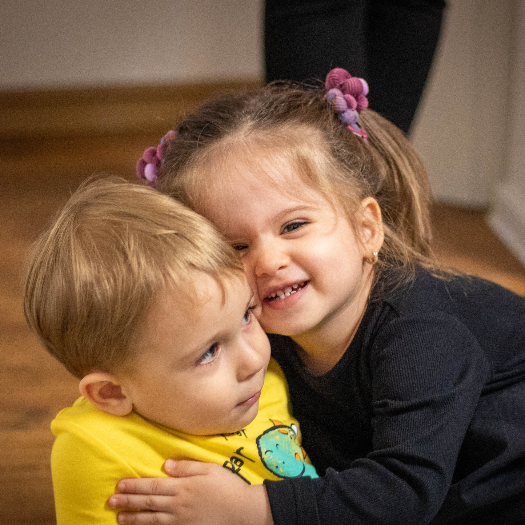 Small child hugging another child with a smile in warm indoor lighting.