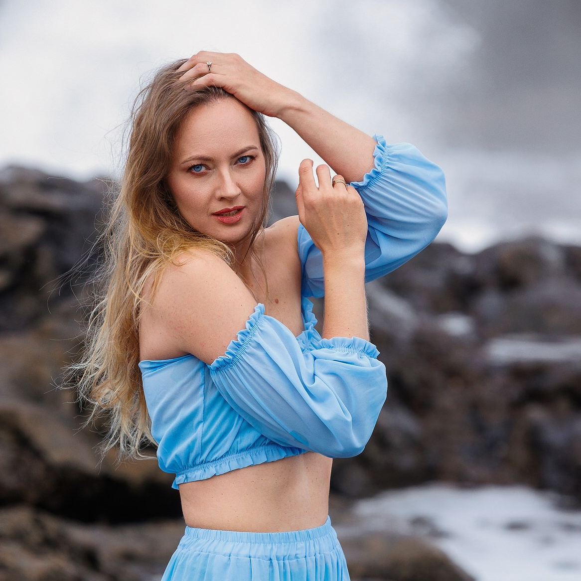 A blonde woman in blue off-the-shoulder top with hands in her hair on rocky coast of Gran Canaria