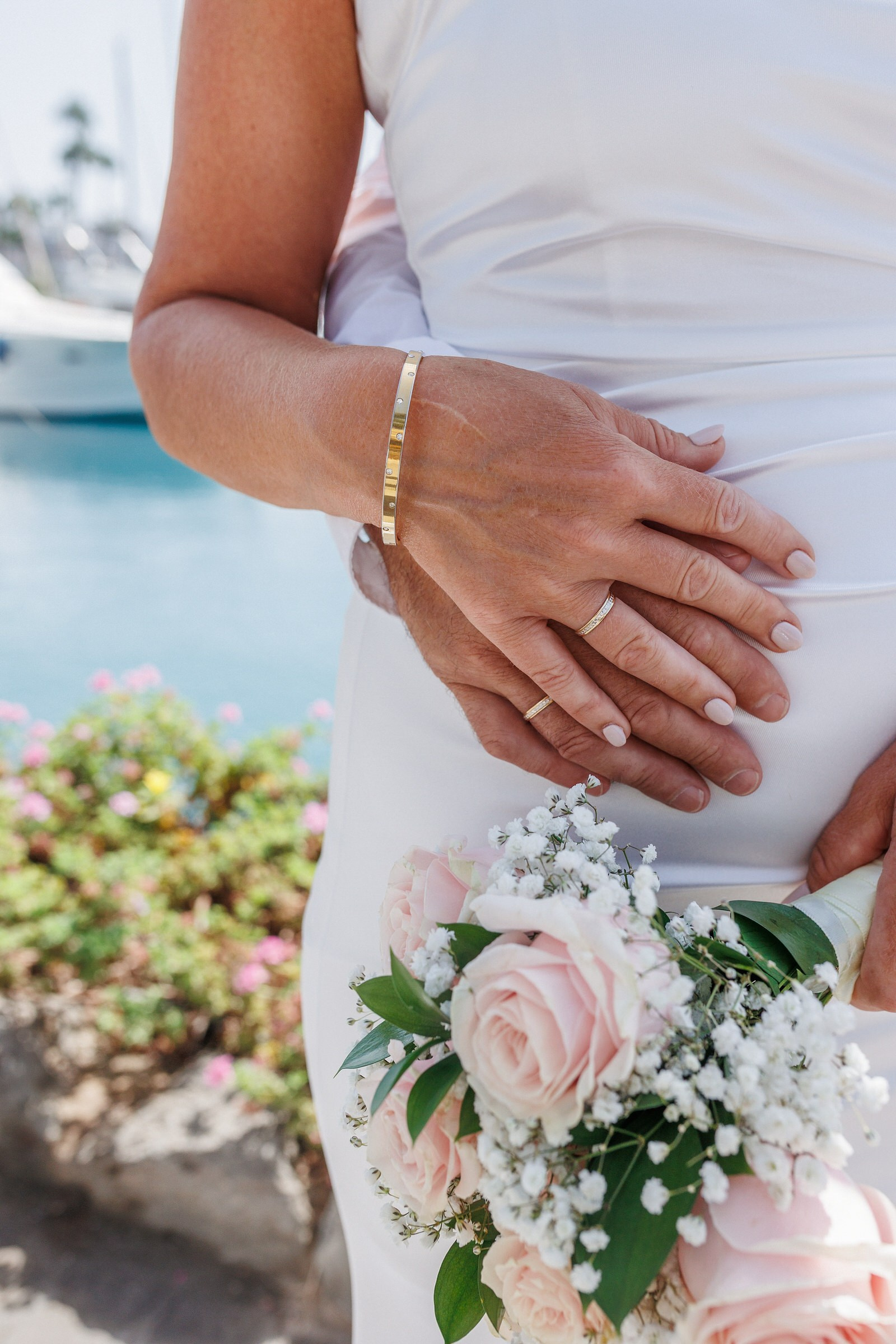 Anfi del Mar: A couple stands close together outdoors near water, with one person holding a bouquet of pink roses and the other resting a hand on their partner’s waist. Gran Canaria