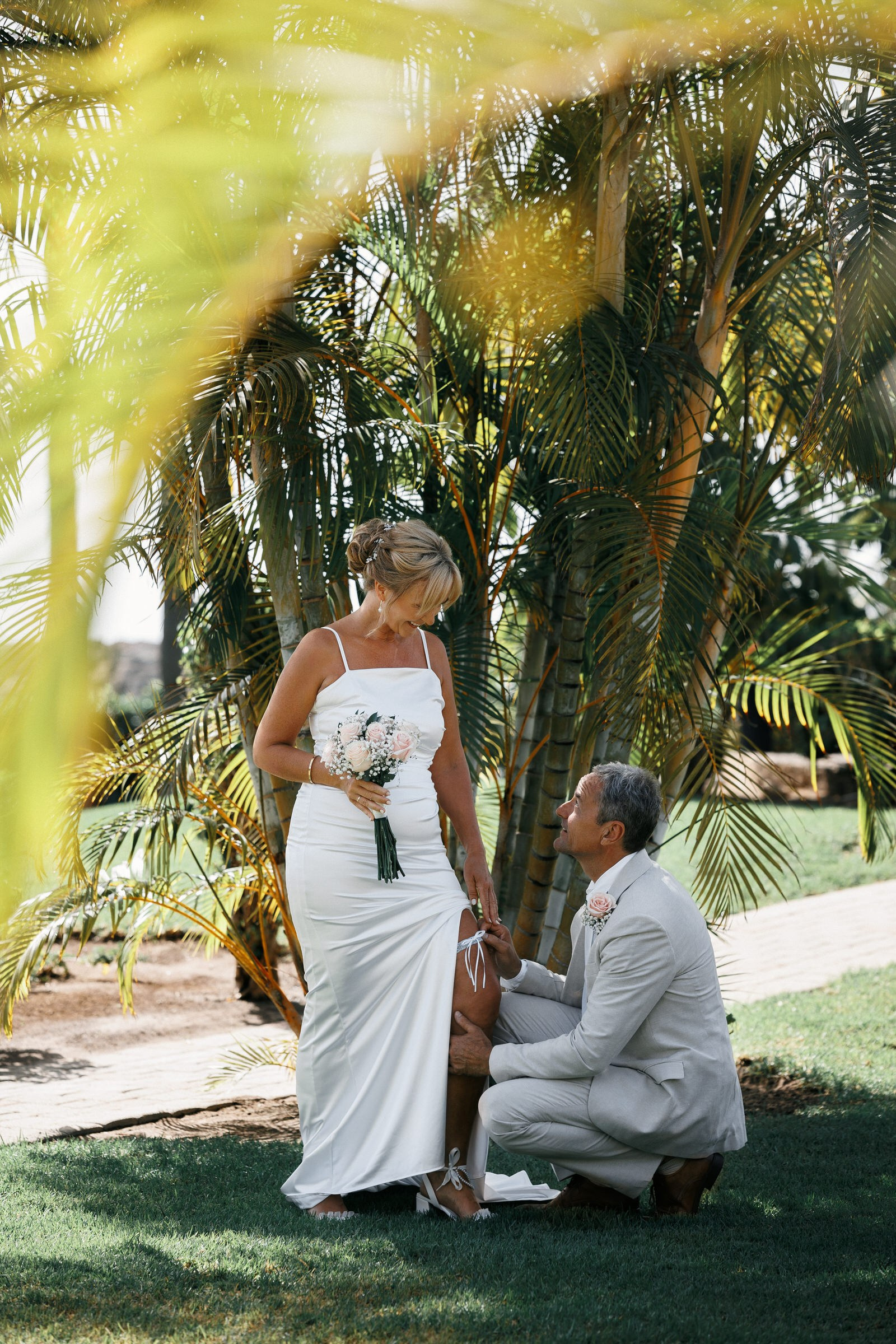 Anfi del Mar: A bride in a white dress stands holding a bouquet while a groom in a light suit kneels to adjust her shoe outdoors near palm trees. Gran Canaria