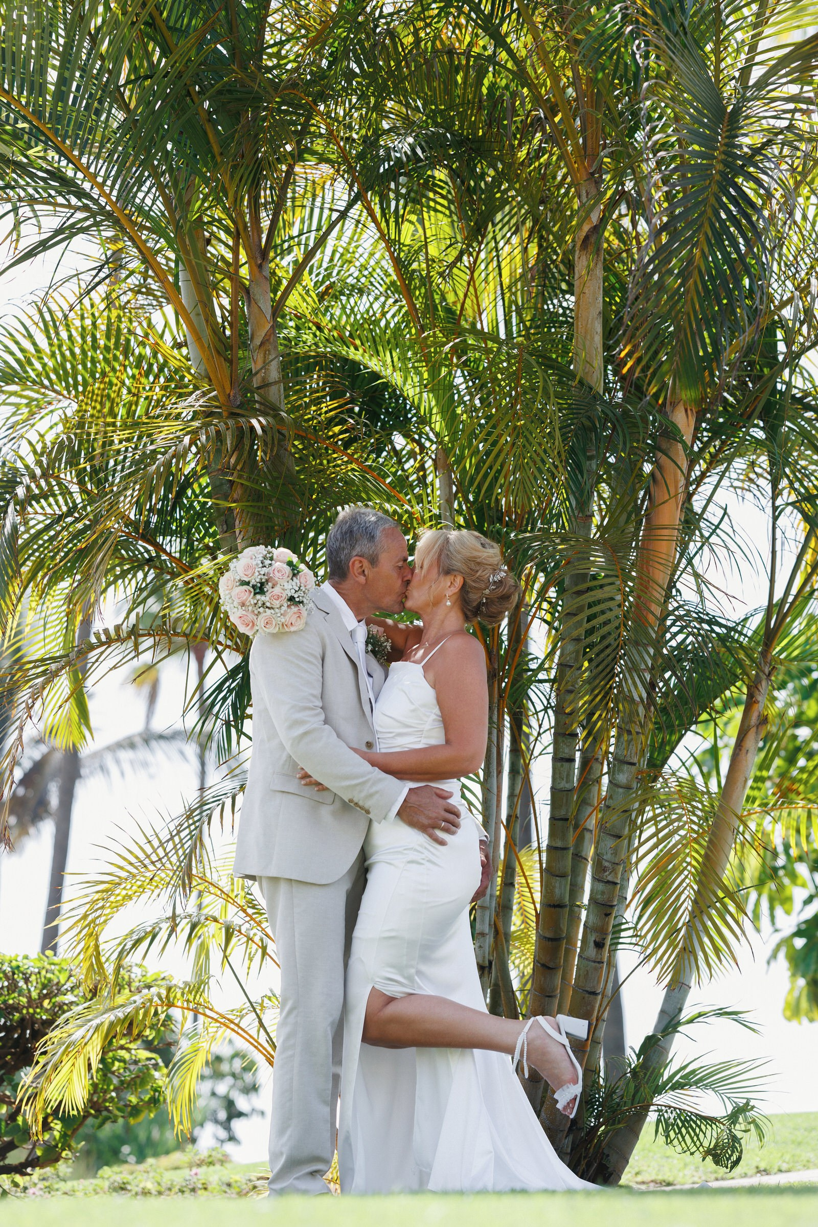 A bride and groom kiss in front of palm trees; the bride holds a bouquet and lifts one leg as the groom embraces her.Gran Canaria