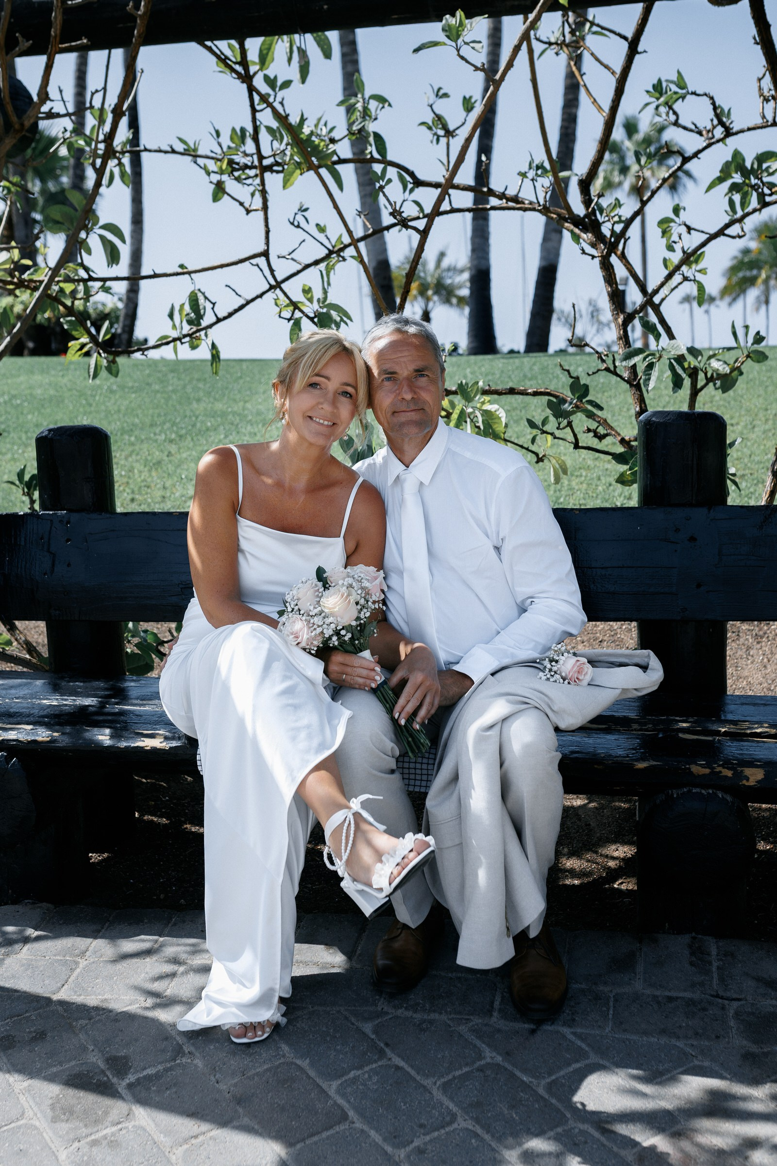 Anfi del Mar: A couple dressed in white formal attire sits on a bench outdoors, holding flowers, with greenery and palm trees in the background.Gran Canaria
