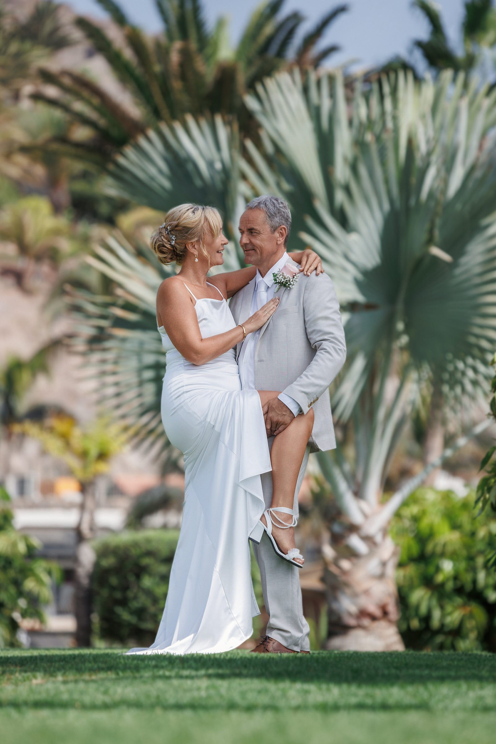 Anfi del Mar: A couple dressed in wedding attire stands outdoors in front of tropical plants, looking at each other and smiling. The woman has her left leg raised, resting it on the man’s knee. Gran Canaria