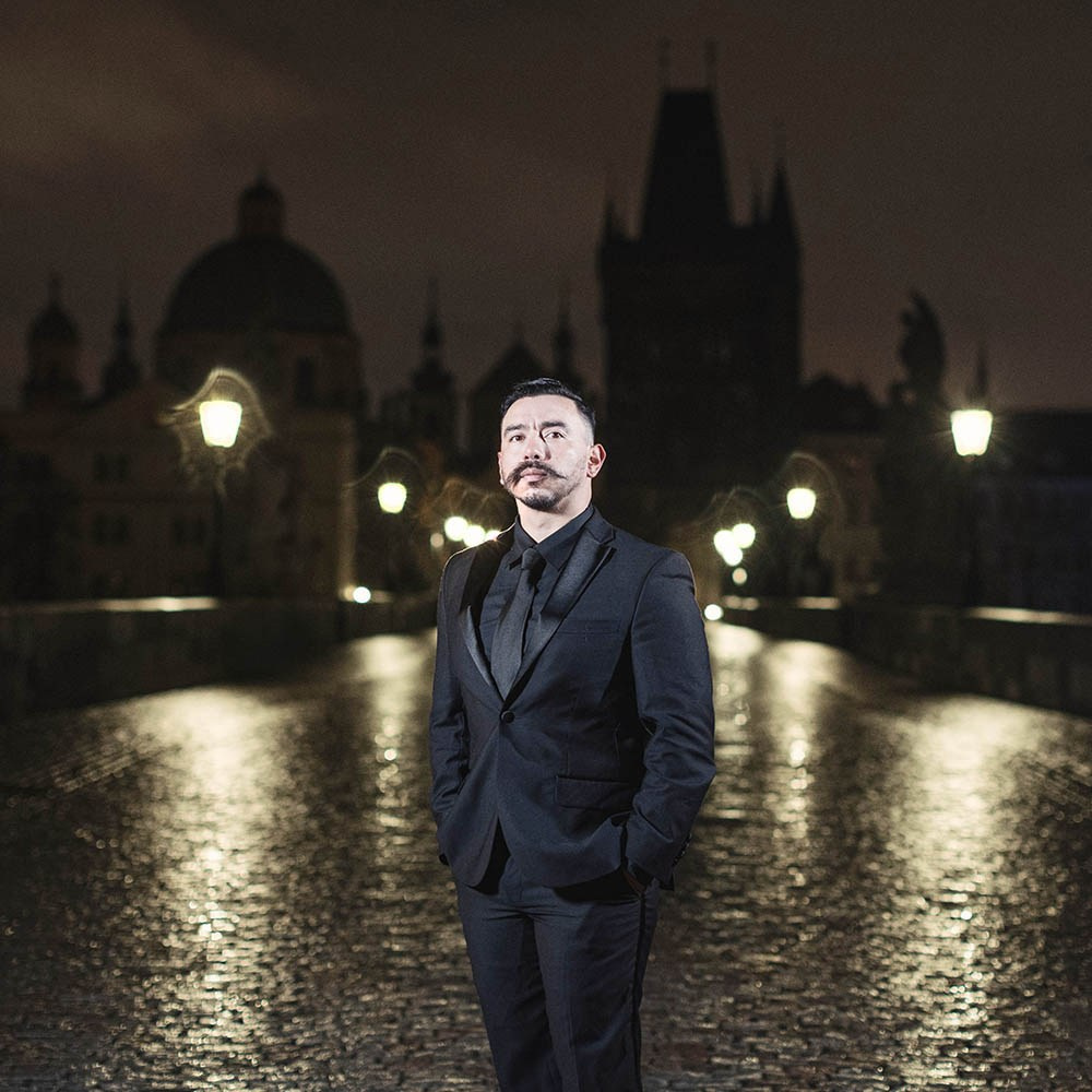The groom, dressed in all black, posing atop the nighttime landscape of a wet Charles Bridge