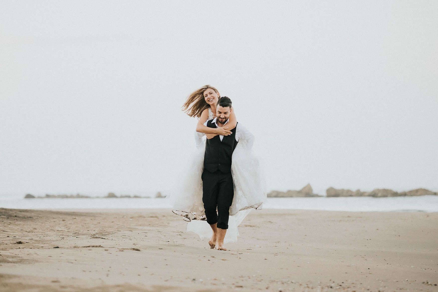 Sposo porta in braccio la sposa lungo la spiaggia, entrambi sorridono liberamente, con lei in un abito da sposa fluttuante e lui in elegante abito nero, in una scena di pura felicità e amore.