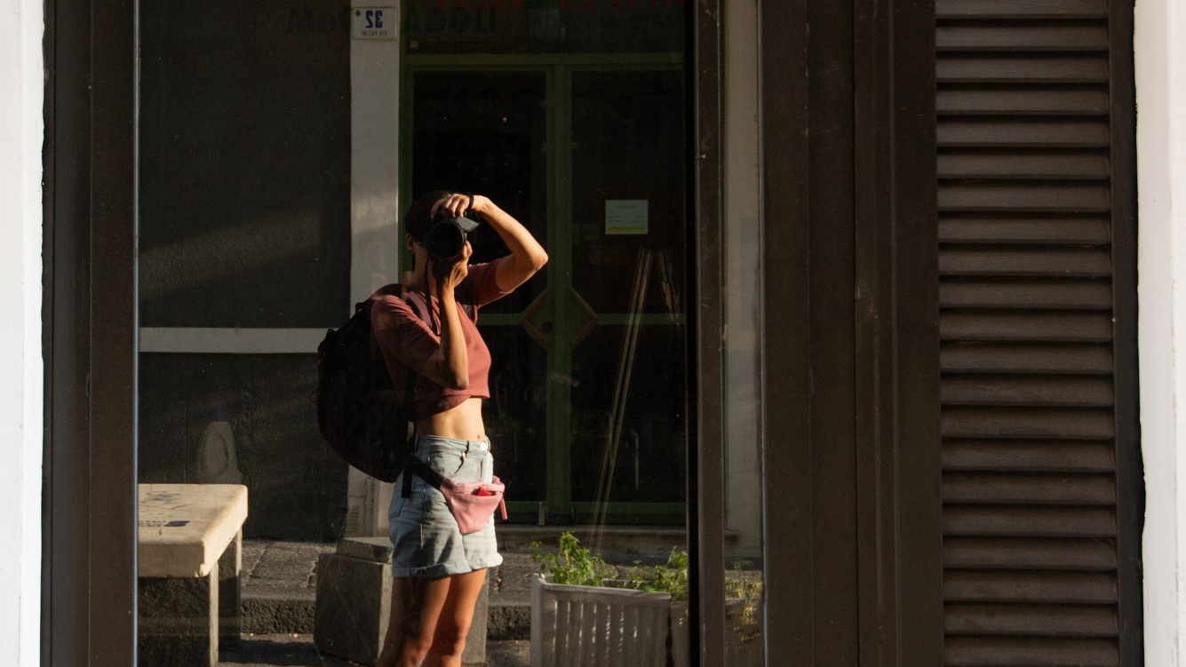 A woman with a backpack photographing her reflection in a glass storefront on a city street