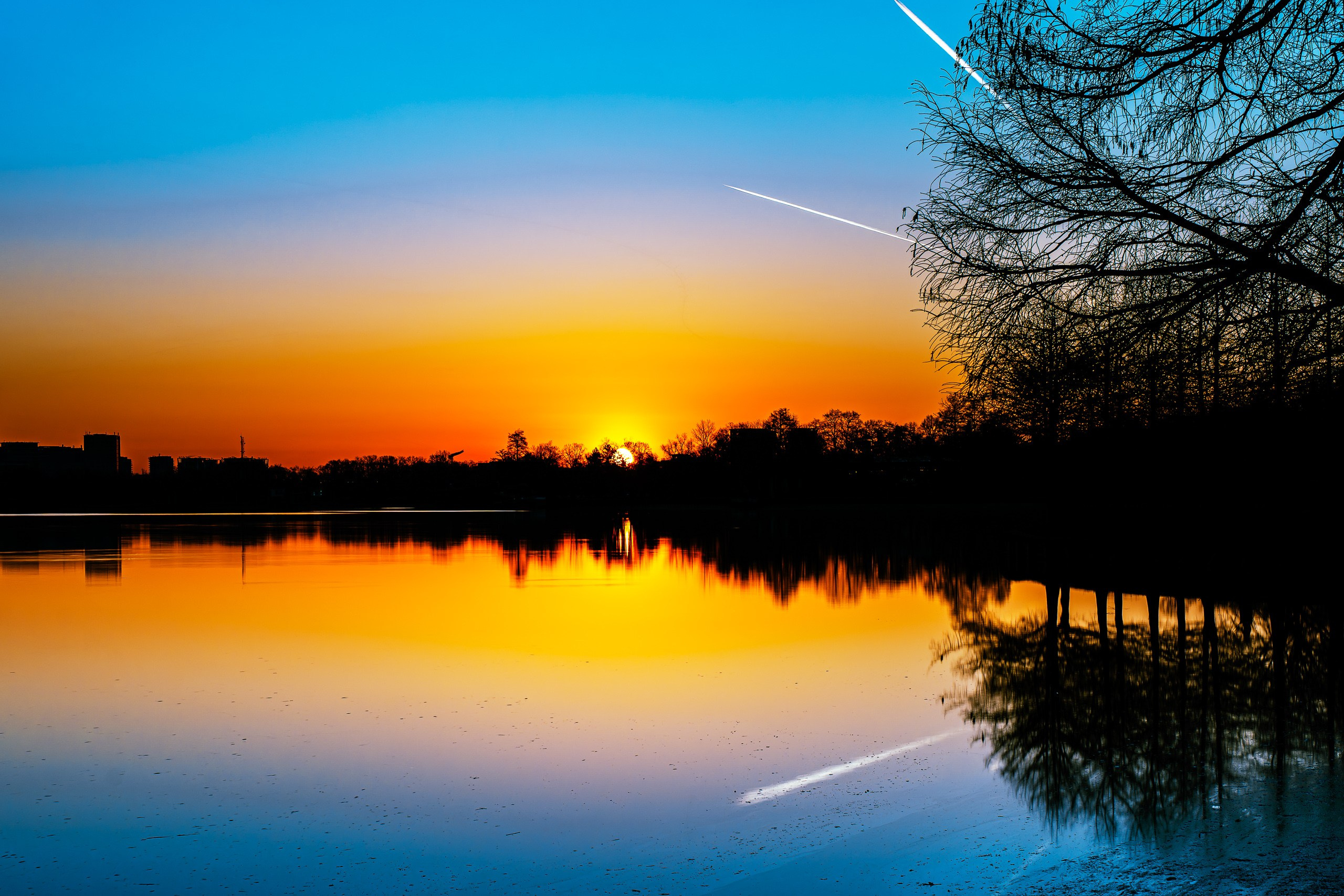 Calm lake at sunset with silhouetted trees and buildings reflected in the water.