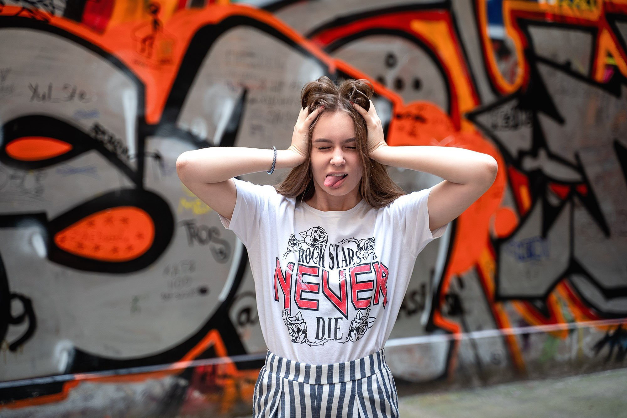 Young woman in shorts and a white t-shirt, playfully covering her ears and eyes while sticking out her tongue, street fashion photo shoot for youth clothing advertisement