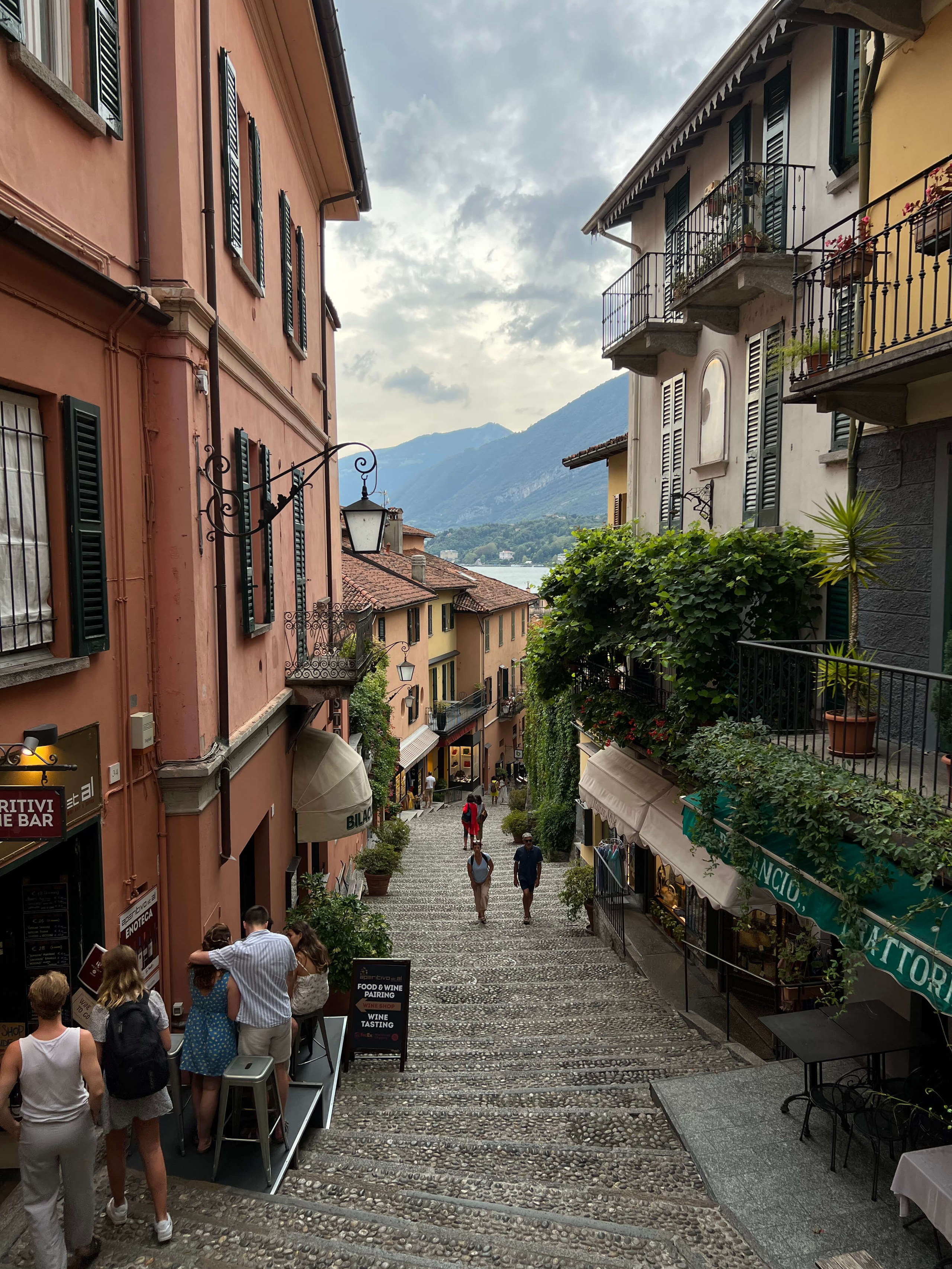 Narrow cobblestone stairway in Bellagio lined with cafés, colourful buildings and Lake Como mountain views
