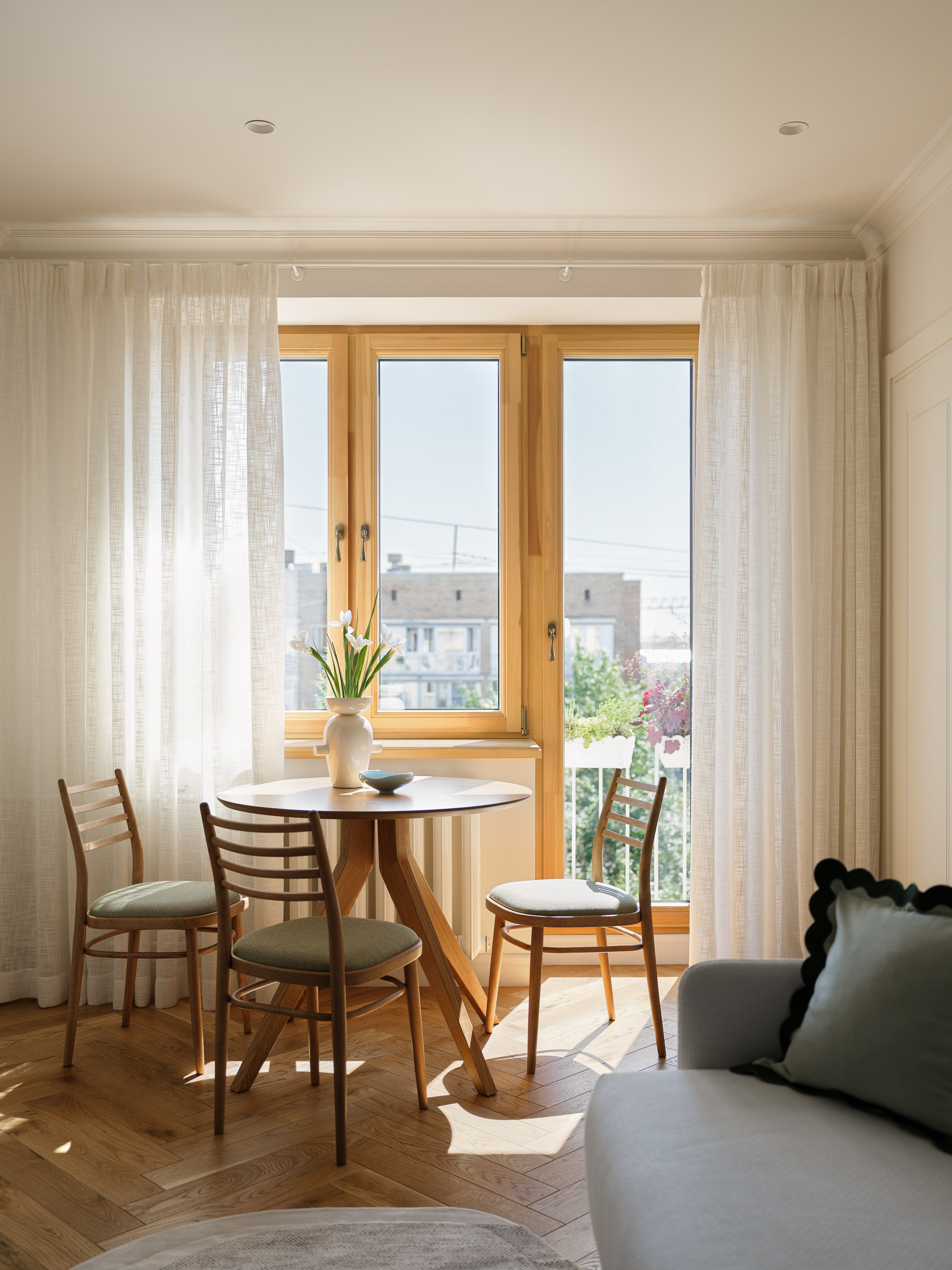 Вид из гостиной сквозь деревянные рамы на зеленый балкон с садовой мебелью.