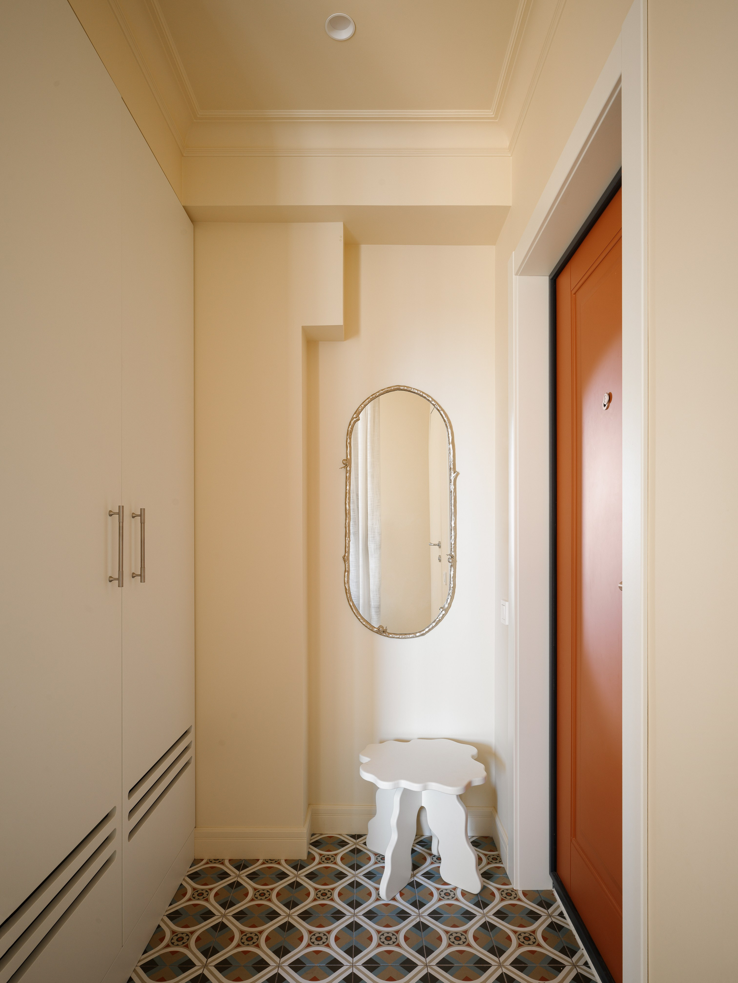 Интерьер прихожей со встроенным шкафом до потолка, мягким пуфом для сидения и настенным зеркалом в овальной раме.