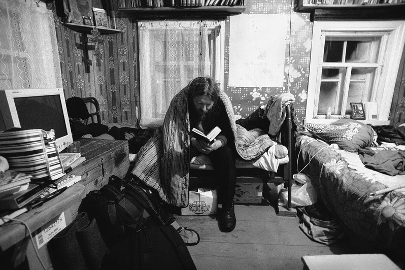 Отец Аркадий, укрывшись ватным матрацем, читает книгу в ещё непротопленном общинном доме, деревня Погост, Пудожский район, Карелия, Россия. Январь 2009