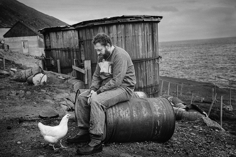Árni Dahl sitting on a barrel and shooting the breeze with the hen, Hattarvik village, Fugloyar municipality, Faroe Islands, Kingdom of Denmark. 1988