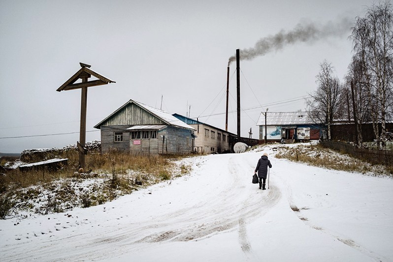 An old woman walks past a roadside cross, Leshukonskoe village, Arkhangelsk region, Russia. October 2017